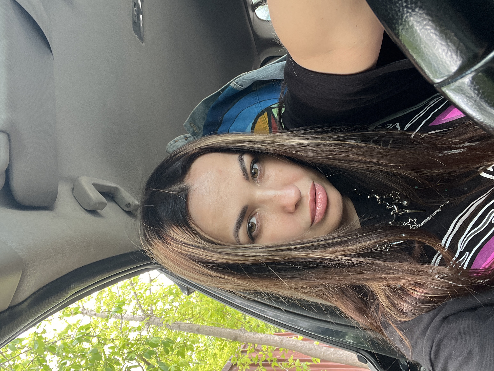A woman is seated inside a car. She has long, brown hair with blonde highlights. She wears a black shirt and a silver necklace. A denim jacket is behind her. Through the car window, green trees and a red fence are visible. The location is Unknown, Unknown.