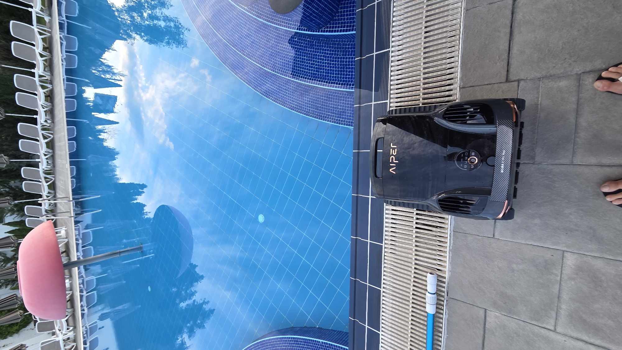 A high-angle shot shows a black pool cleaning robot with the brand name "AIPER" printed on its top, sitting on a tiled patio beside a swimming pool. The robot is positioned near a slatted drainage grate that runs along the edge of the pool. The pool itself is a vibrant blue, with tiled steps visible on the right side. The water reflects the sky and surrounding trees. In the background, a row of lounge chairs and umbrellas are set up next to the pool. On the left side of the pool, a pink umbrella-shaped structure juts out of the water. The overall scene suggests a resort or hotel pool area.