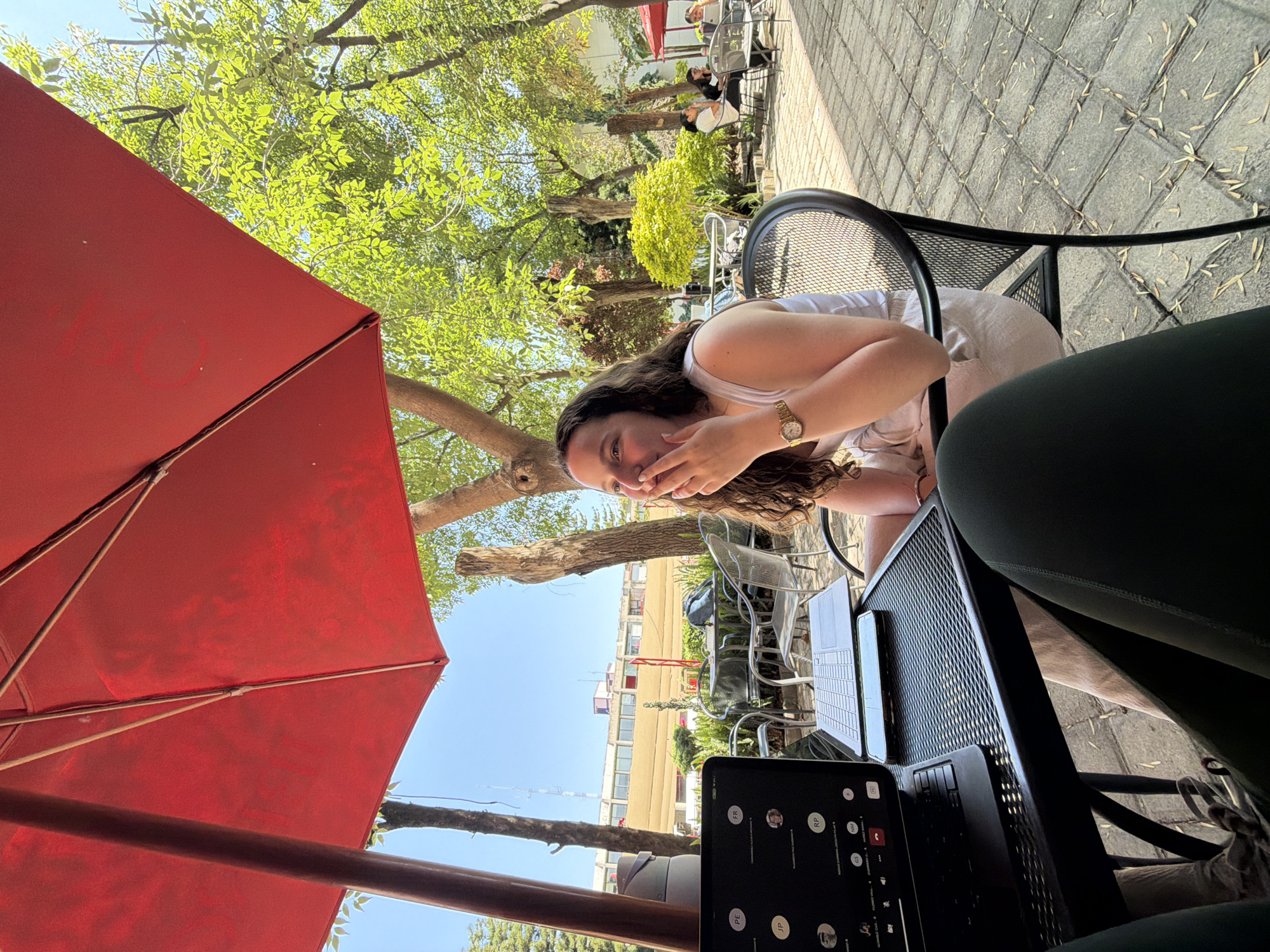 The image captures an outdoor café or patio scene under bright daylight. In the foreground, a woman with long, curly brown hair, wearing a white tank top and a gold watch, is seated at a black metal outdoor table. She is looking towards the camera, covering her mouth with her hand, suggesting amusement or a reaction to a comment. Her leg, dressed in dark green leggings, is partially visible in the bottom right corner.
On the table in front of her, multiple electronic devices are present: a laptop with an illuminated keyboard, a smartphone, and a tablet displaying what appears to be a video call interface. The tablet screen shows circular profile pictures and initials such as "FR," "RP," "GS," "PE," and "JP," indicating participants in a virtual meeting or chat. Another laptop is also visible further back on the table.
The setting is an outdoor area, heavily shaded by a large red patio umbrella that dominates the left side of the frame. Lush green trees are abundant, casting dappled light and shadow across the scene. The ground is paved with grey stones in a grid pattern, with scattered dried leaves. In the mid-ground, other individuals are seated at similar outdoor tables, some partially obscured by trees and shadows. In the distance, under a clear blue sky, a line of multi-story buildings is visible, suggesting an urban or suburban environment. The scene depicts a casual, bright daytime gathering, possibly involving work or study alongside social interaction.