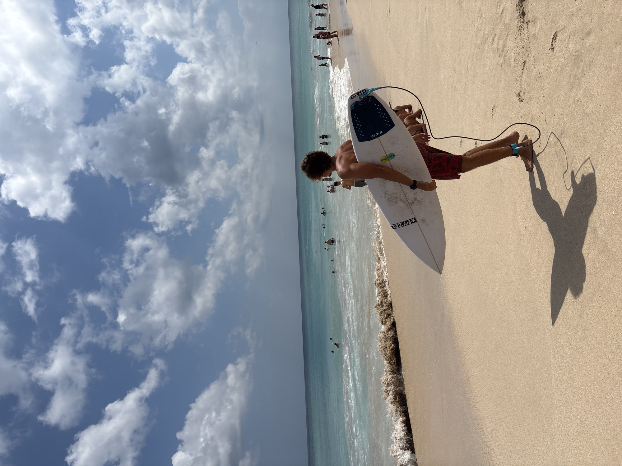 The image shows a surfer on a sandy beach carrying a surfboard. The surfer, seen from behind, is wearing red patterned swim trunks and has a black ankle leash attached to the surfboard. The surfboard is white with a blue and black grip pad at the tail. In the background, the turquoise ocean meets the shore with gentle waves. Several people are visible in the water, swimming and enjoying the beach. The sky is blue with scattered white clouds. The scene appears to be a sunny day at a beach, likely a popular surf spot. While the specific location is not identifiable from the image alone, Kuta Selatan, Indonesia is a well-known area for surfing, with beaches like Uluwatu and Padang Padang attracting surfers.