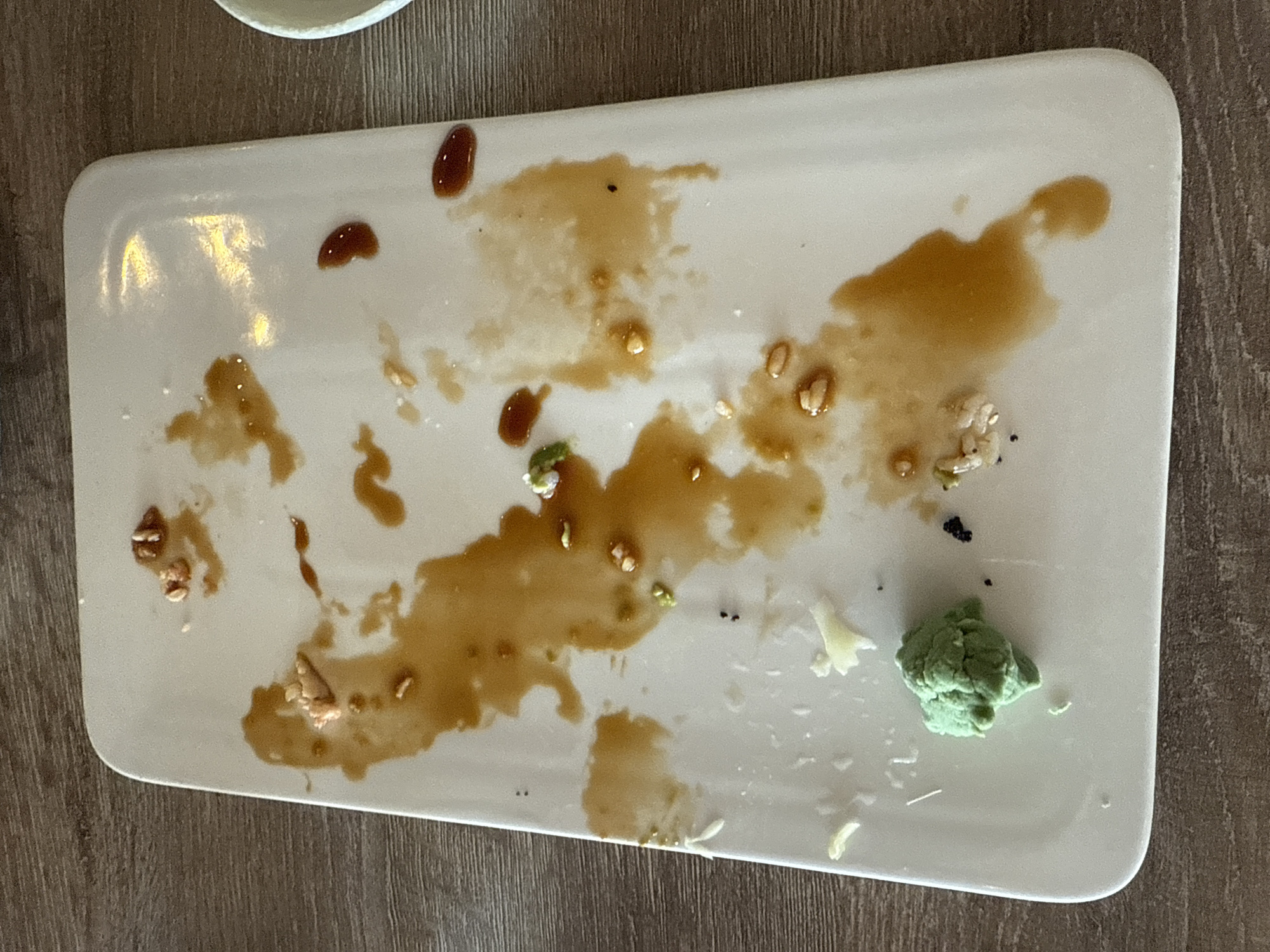 The image shows a rectangular white plate, likely ceramic, containing remnants of food. There are visible traces of a dark brown sauce, possibly soy sauce or teriyaki sauce, along with scattered small food particles. A ball of green wasabi is located in the lower right corner of the plate. The plate is positioned on a wooden surface. The location is possibly Darien, United States of America. There are no people in the image.