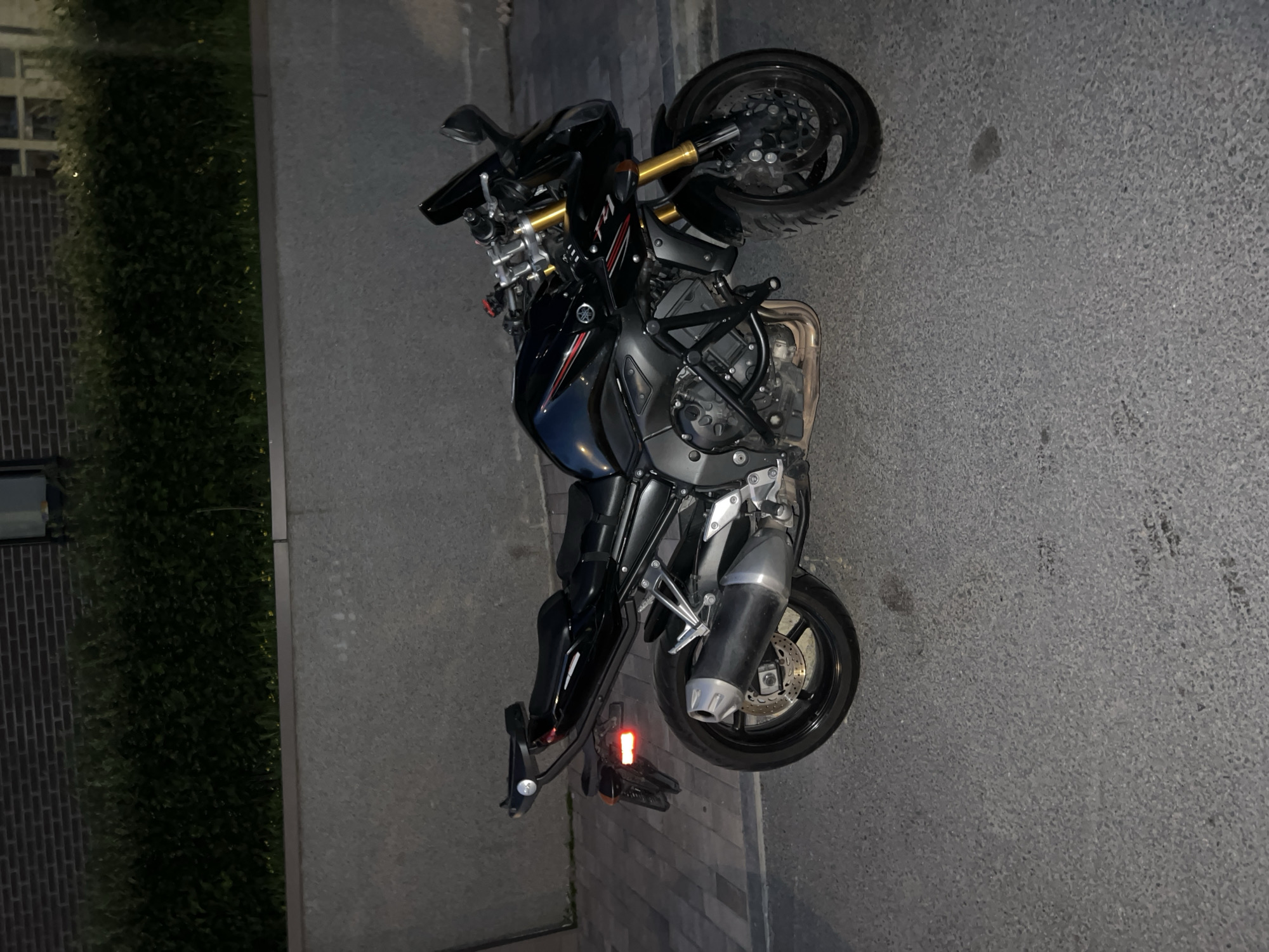 A black Yamaha FZS motorcycle is parked on an asphalt road at street level. The motorcycle is facing right, with its kickstand down. The front of the motorcycle is slightly obscured by a dark, angular object. The motorcycle has a black seat, a black fuel tank with red and white accents, a black exhaust pipe, and gold-colored front forks. A black metal frame is attached to the motorcycle's engine. In the background, a textured gray wall runs horizontally, with a dark green hedge above it. A brick building is visible behind the hedge, with a window on the right side. The ground in front of the wall is paved with gray interlocking stones. The lighting suggests it is nighttime or dusk, with artificial light casting faint shadows. There are no people visible in the image.