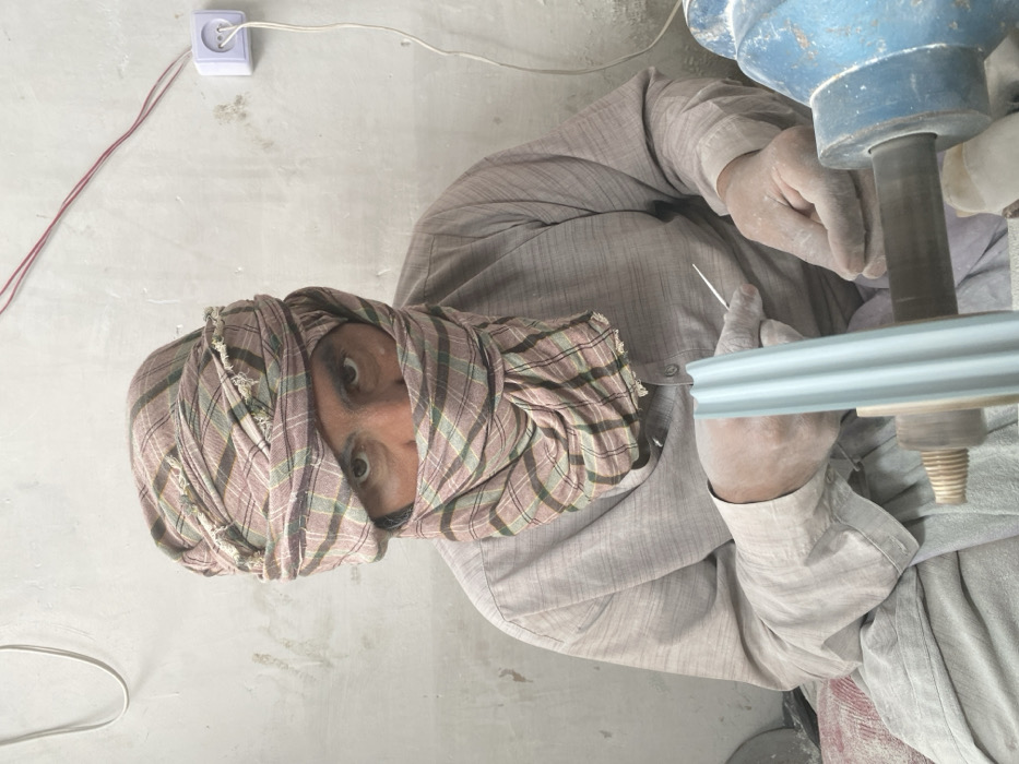An adult male is observed in a workshop in Kandahar, Afghanistan, engaged in a manufacturing or crafting process. His face is largely obscured by a plaid head covering, revealing only his eyes, which look directly towards the viewer. He wears a light-colored, long-sleeved garment, possibly a traditional shalwar kameez top. His hands are dusted with a light powder. He holds a thin, pointed metal tool in his right hand. A light blue, fluted cylindrical object is positioned vertically on a wooden spindle, which is connected to a partially visible blue machine on the right, suggesting a lathe or polishing apparatus. The background consists of a plain, light-colored wall with exposed white and red electrical wiring. A white electrical outlet with a plugged-in white cord is visible on the upper right wall. The scene depicts an active workspace with tools and materials.