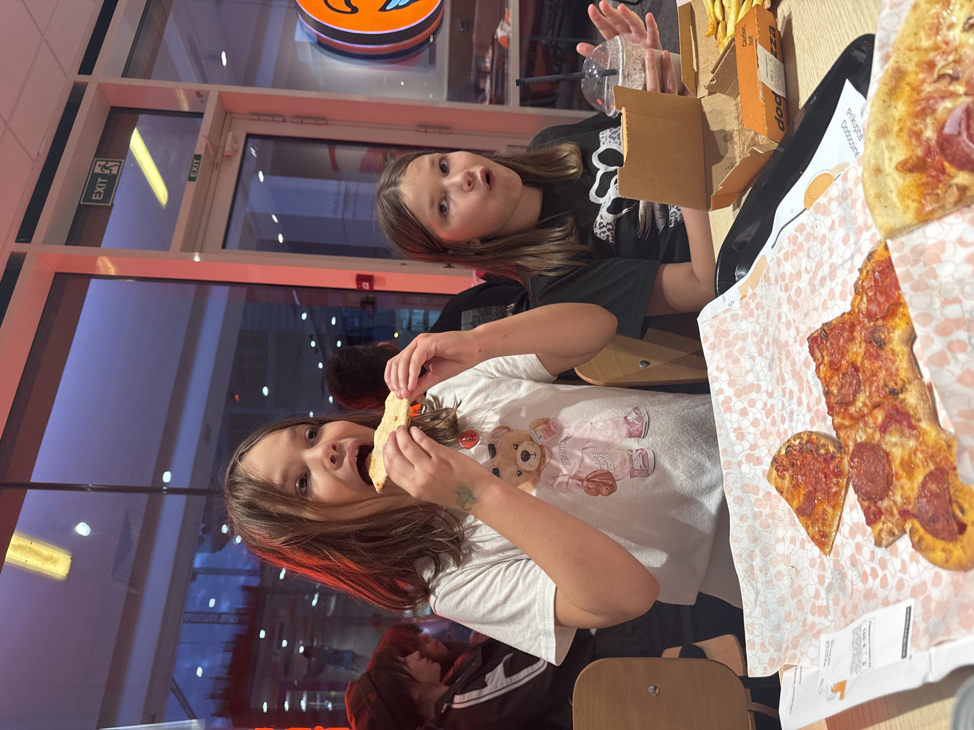 Two young girls are seated at a table in what appears to be a casual restaurant or pizzeria, possibly in Budva, Montenegro. The scene is brightly lit by artificial light, suggesting it could be day or night.
The girl on the left, wearing a black t-shirt, looks towards the camera with a wide-eyed, slightly open-mouthed expression, holding a clear plastic cup with a straw. Her long brown hair falls over her shoulders. The girl on the right, younger and wearing a white t-shirt with a teddy bear design that reads "A HAPPY LIFE," is actively taking a bite from a slice of pizza, also looking towards the camera with an open mouth and an excited expression. Her long brown hair is also prominent.
On the table in front of them are several items of food: slices of pizza, some with pepperoni, laid out on patterned paper liners, and French fries in an open cardboard box. A black tray is also visible. The restaurant features light-colored walls, large glass windows or doors, and orange accents on the walls and ceiling.
Visible text includes "EXIT" on green signs, "dog pizza" on the cardboard box holding fries, and "Prikupite Dodoccoine" (meaning "Collect Dodoccoins" in Croatian/Serbian, hinting at a loyalty program) printed on the pizza paper liner. The overall atmosphere is lively and cheerful, with both girls seemingly enjoying their meal and each other's company.