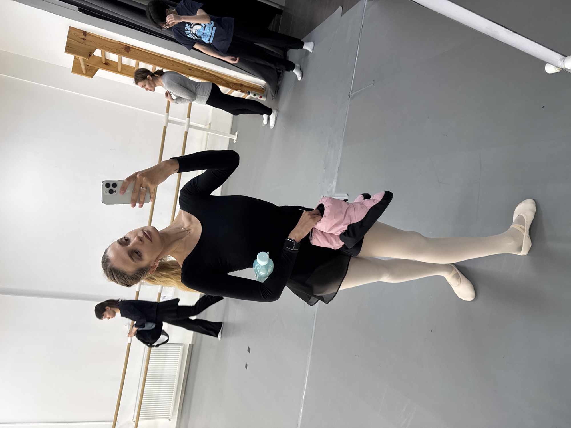 This indoor photograph captures a dance studio in Tbilisi, Georgia, with a woman taking a selfie in a large mirror. The main subject, positioned prominently, has long blonde hair tied back and is dressed in a black long-sleeved leotard, a sheer black skirt, light-colored ballet tights, and white ballet slippers. She holds a light-colored smartphone in her left hand, directing it towards the mirror to take her picture, and looks at the phone's screen. A light blue water bottle is visible tucked into her waist, and she holds a pink fabric item in her right hand. She wears a watch on her left wrist and a ring on her left ring finger.
The mirror reflects other individuals and studio features. In the mid-ground, a woman with dark hair, wearing a grey long-sleeved top and dark pants with white socks, stands near a wooden ballet barre. Further back, another person in a dark blue t-shirt with a white graphic that appears to say "ICE COLD" or "COOL OFF," along with dark pants and white socks, is also reflected. Closer to the main subject, another woman in black clothing and carrying a black backpack is partially visible.
The setting includes white walls, a grey floor, and multiple wooden ballet barres. A white radiator is also visible in the background reflection. The lighting is bright and consistent, indicating daytime. The scene depicts a moment during or between dance practice sessions.