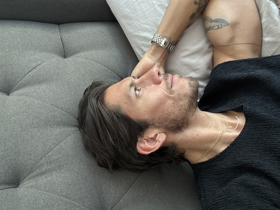 A man reclines on a grey, button-tufted couch with his head resting on a white pillow. He wears a black textured shirt and two delicate gold necklaces. His dark hair is slightly disheveled, and he has light stubble on his face. A silver watch adorns his wrist. Tattoos are visible on his upper arm.