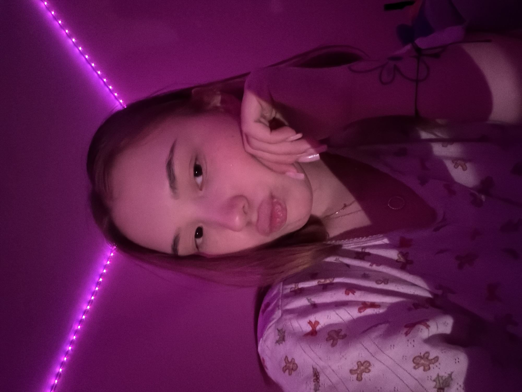 A young woman poses for a selfie in a room illuminated by purple LED strip lights. She rests her face on her hand, puckering her lips. She wears a patterned top, possibly pajamas, with small gingerbread man-like figures. A plush toy is visible in the background. There is no information to determine if the location is in the city of Chaikovsky, Russia.
