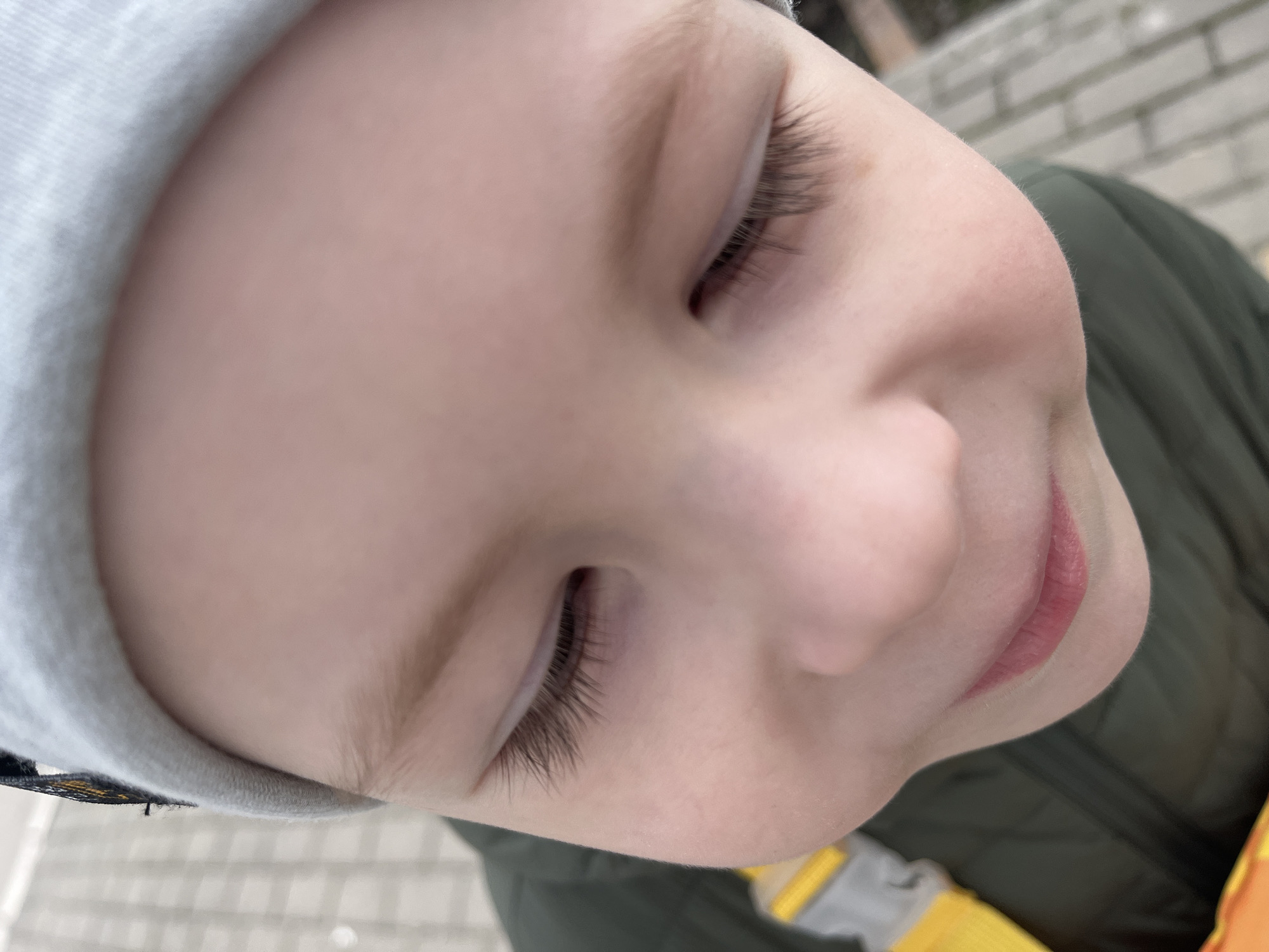 A close-up, slightly tilted shot focuses on a child's face. The child, wearing a light-colored knit hat, has fair skin, closed eyes with visible eyelashes, and a gentle smile. Part of the child's upper body is visible, clad in an olive green jacket with a hint of yellow underneath. The background is blurred, suggesting an outdoor setting. In the periphery, parts of the paving stones are visible, as well as the wall. The location is identified as Usman, Russia.