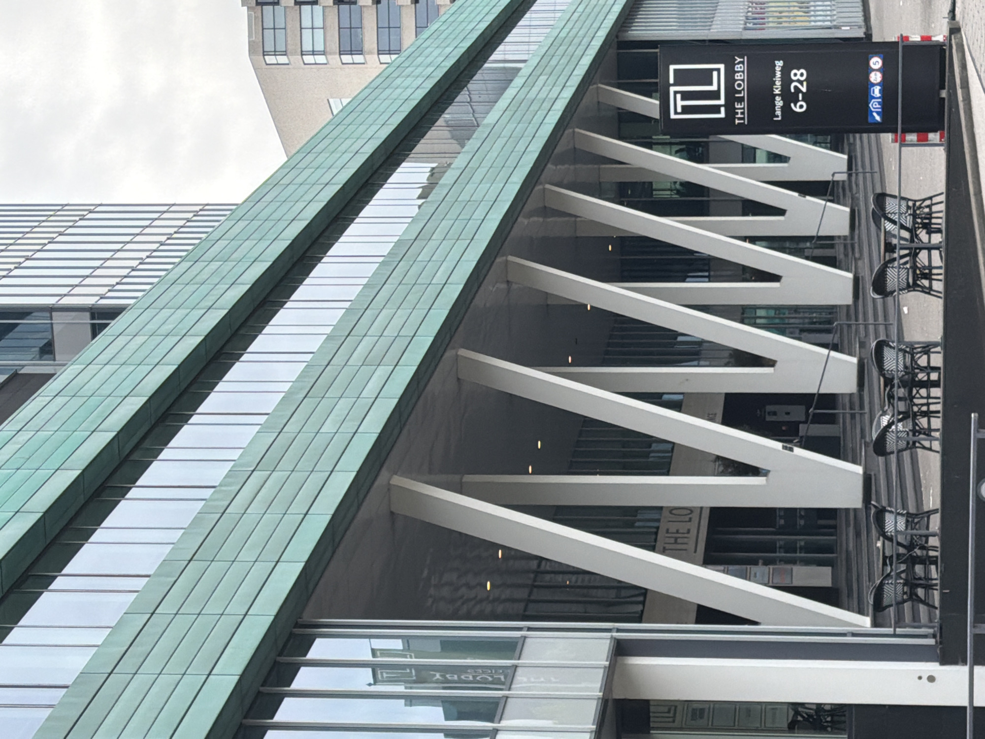 The image shows a modern building with a distinctive green facade composed of horizontal panels and numerous vertical windows. The lower level of the building features a covered walkway supported by angled white columns. In front of the building, several black wicker chairs and tables are arranged on a patio. To the right, a black signpost displays the text "TL THE LOBBY Lange Kleiweg 6-28" along with various parking and traffic symbols. The scene is set in a city environment, indicated by the architecture and the presence of street furniture. Based on the address displayed on the signpost, the location is Lange Kleiweg 6-28 in Rijswijk, Netherlands. There are no people visible in the image. The sky is overcast.