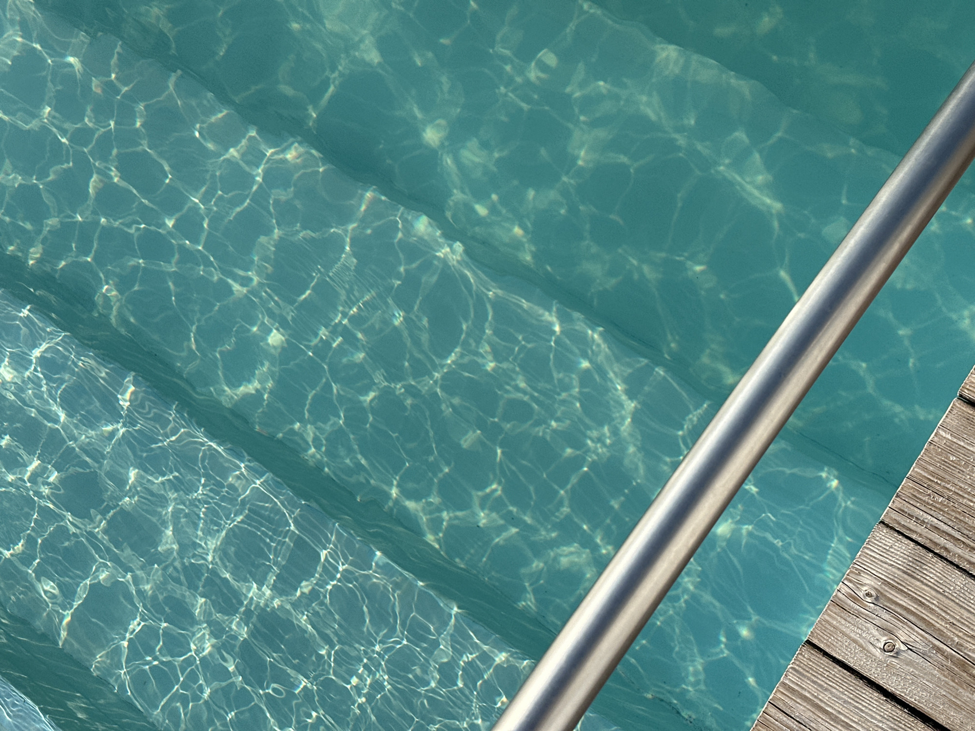 A close-up, high-angle shot captures the surface of a swimming pool with distinct, dappled light patterns reflecting off the turquoise water. Visible beneath the water are several steps, also appearing turquoise. A portion of a wooden deck runs diagonally across the bottom left of the frame, with a smooth, silver-toned metal handrail extending from the bottom right towards the center. The image does not contain any individuals. This scene is in Yaroslavskaya, Russia.