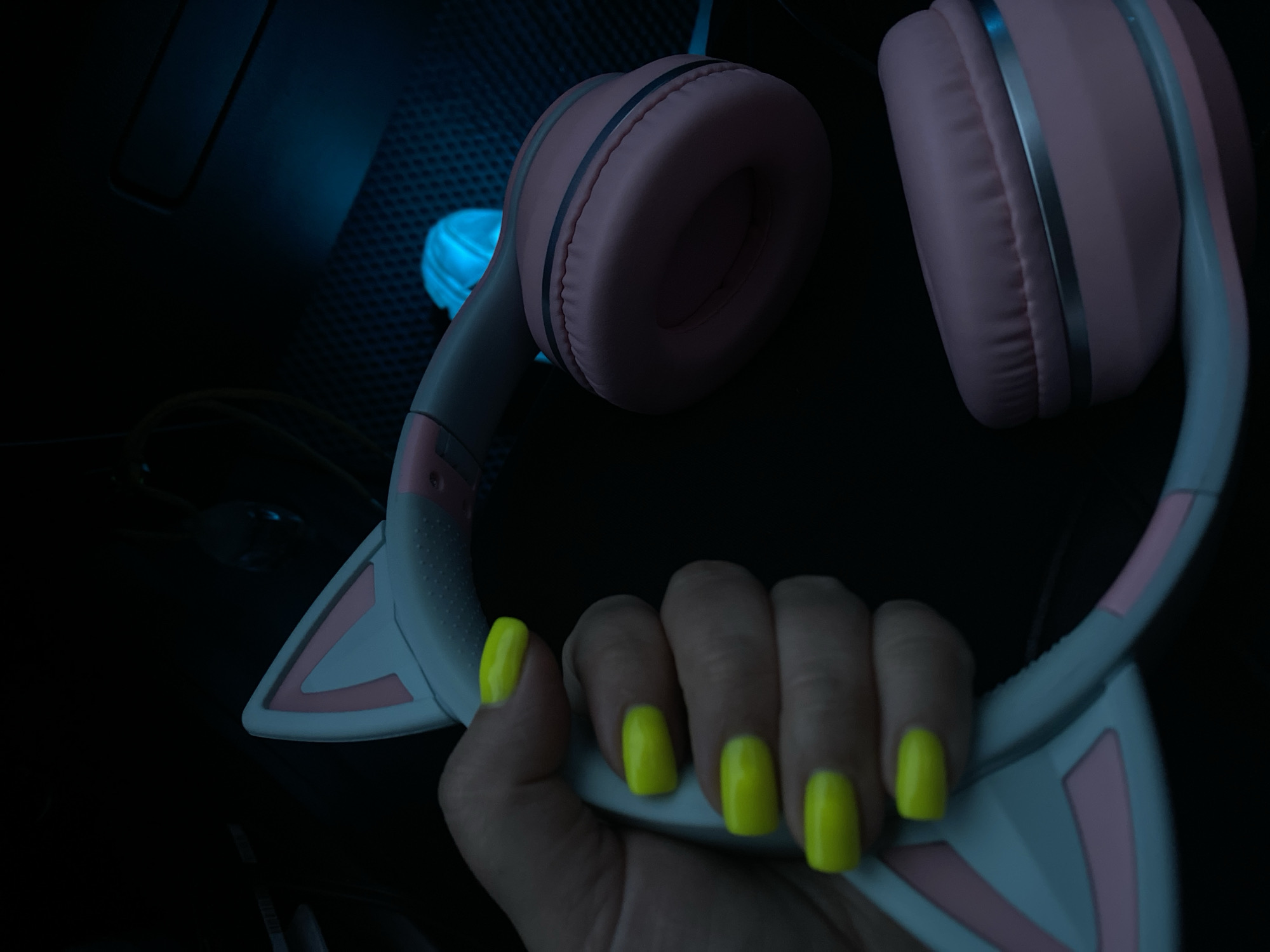 A hand with bright yellow fingernails holds a pair of pink and white cat-ear headphones. The headphones are resting on a dark, textured surface that appears to be a laptop. In the background, a blue light emanates from the laptop, casting shadows and creating a moody atmosphere. The overall scene suggests a personal space, possibly a bedroom or a gaming setup, in a home in Balashikha, Russia.