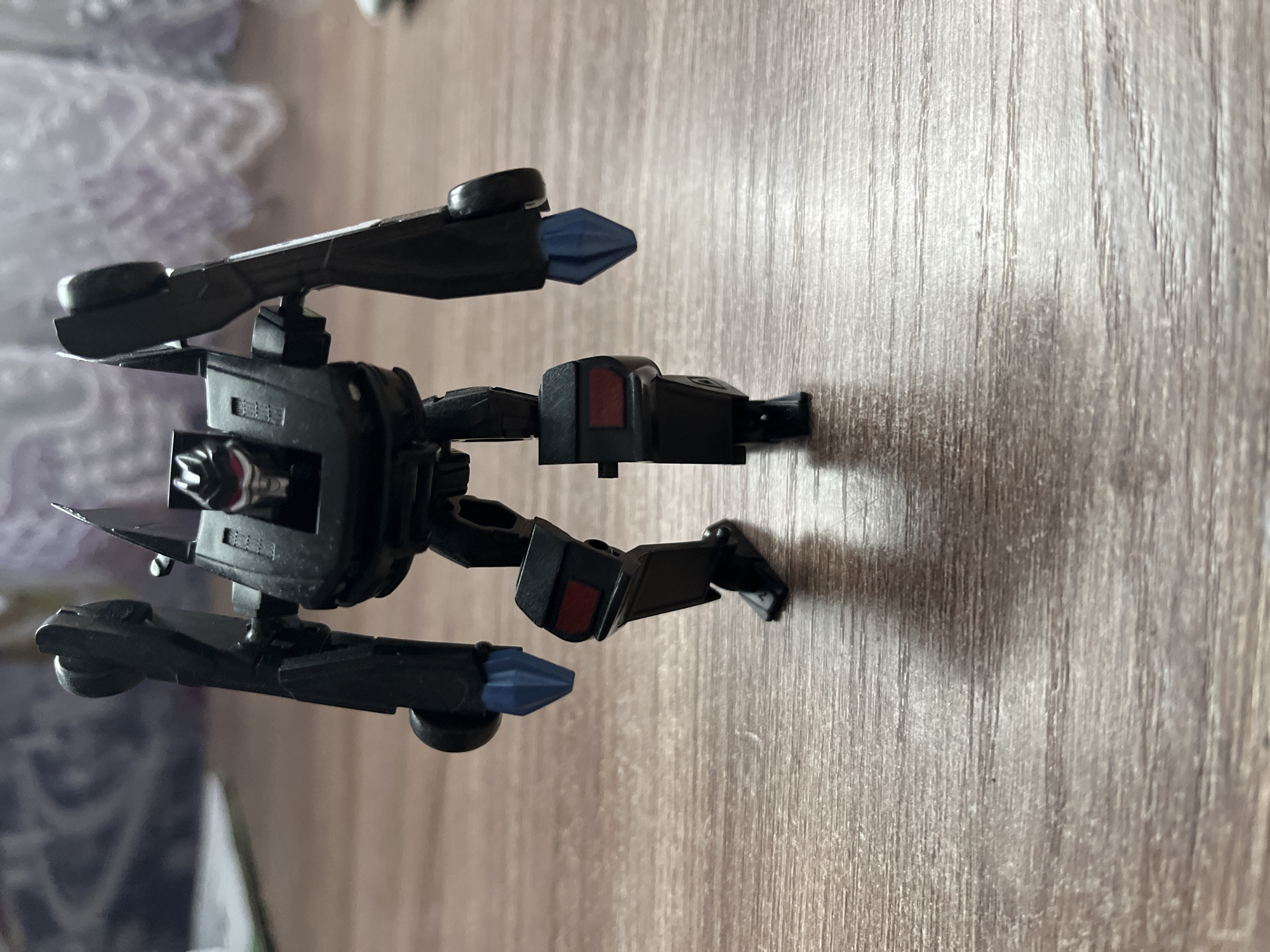 A black, plastic toy robot is on a light brown, wood-grain surface. The robot has a central body with arms and legs extending outwards. The ends of the arms and legs have blue, pointed tips. The background includes a blurry, light purple and white patterned fabric. The location is Марковский, Russia.