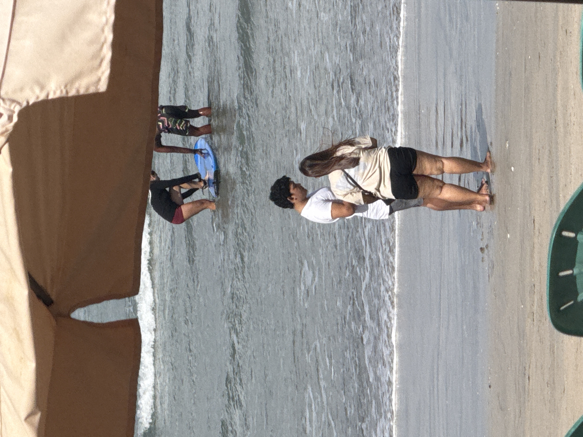 A group of people are visible on a beach in Kuta, Indonesia, in front of a body of water. In the foreground, a couple stands on the wet sand, facing each other. The man is wearing a white t-shirt and dark shorts, and the woman is wearing a tan t-shirt and black shorts. Further back, two individuals are in the water, one on a blue surfboard, and another standing nearby. The sea is a calm gray-blue, with small waves breaking near the shore. The scene is framed by a large, tan canopy from above.