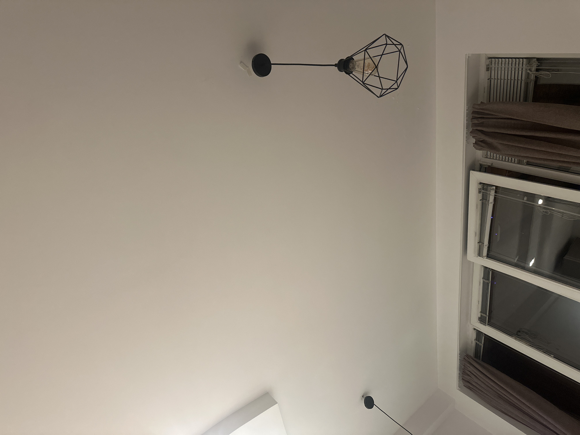 A low-angle shot reveals a white ceiling with two pendant lights and a window, possibly in Sofia, Bulgaria. The pendant lights feature black fixtures, with one incorporating a geometric, cage-like lampshade. The window is open, revealing curtains and the outside view. The overall composition is minimalist and emphasizes the ceiling and lighting elements.