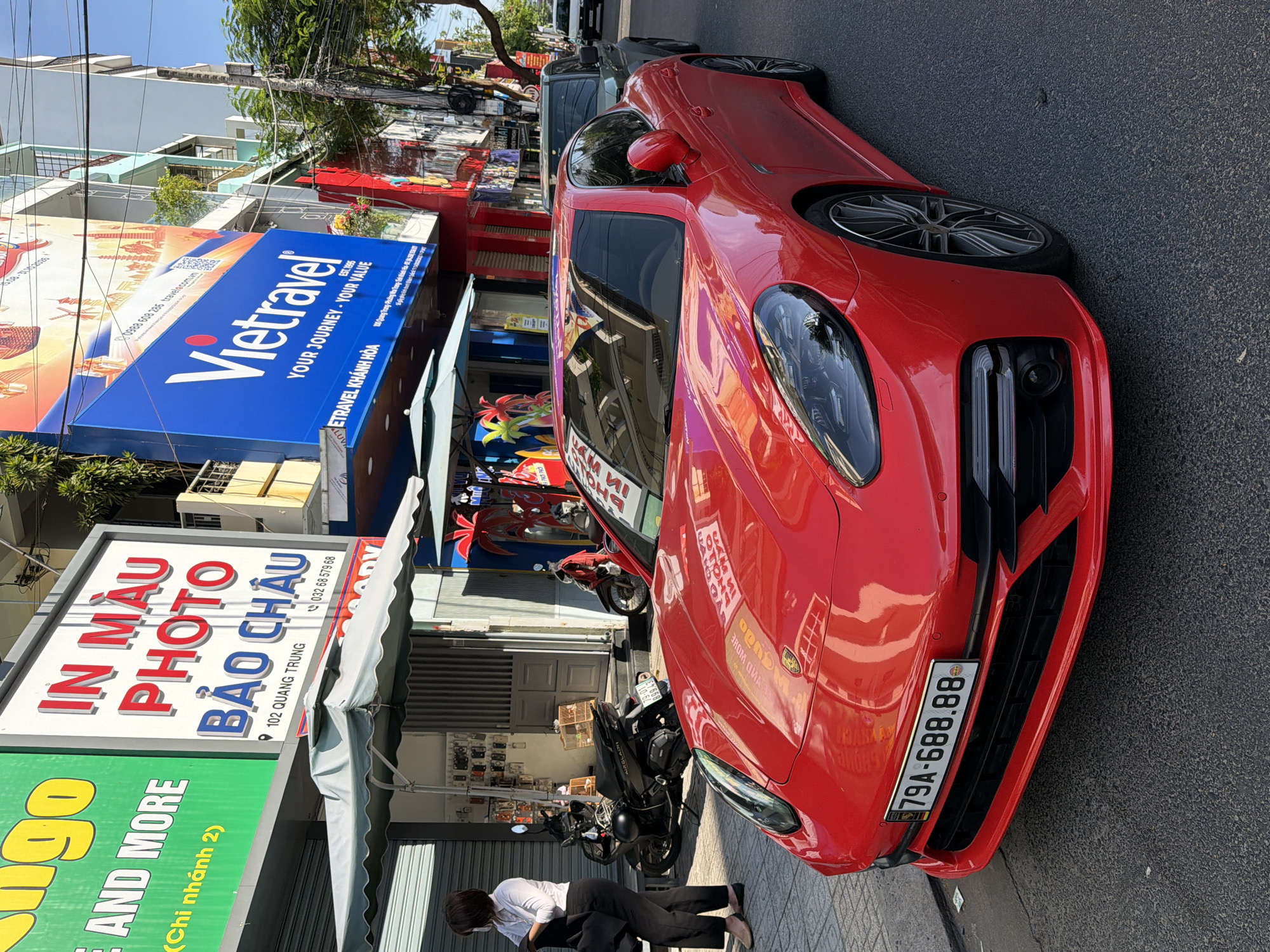 A bright red Porsche Panamera is parked on the side of a street in Nha Trang, Vietnam. The car is the primary subject, occupying the majority of the frame. Its license plate reads "79A-688.88". In the background, buildings with signs are visible, including one for "Vietravel" and another for a photo shop. A woman in black pants and a white shirt stands on the sidewalk, holding a black jacket. Motorcycles are parked near the buildings. The street appears to be paved, and the weather seems to be sunny. The overall scene suggests an urban environment in Vietnam.