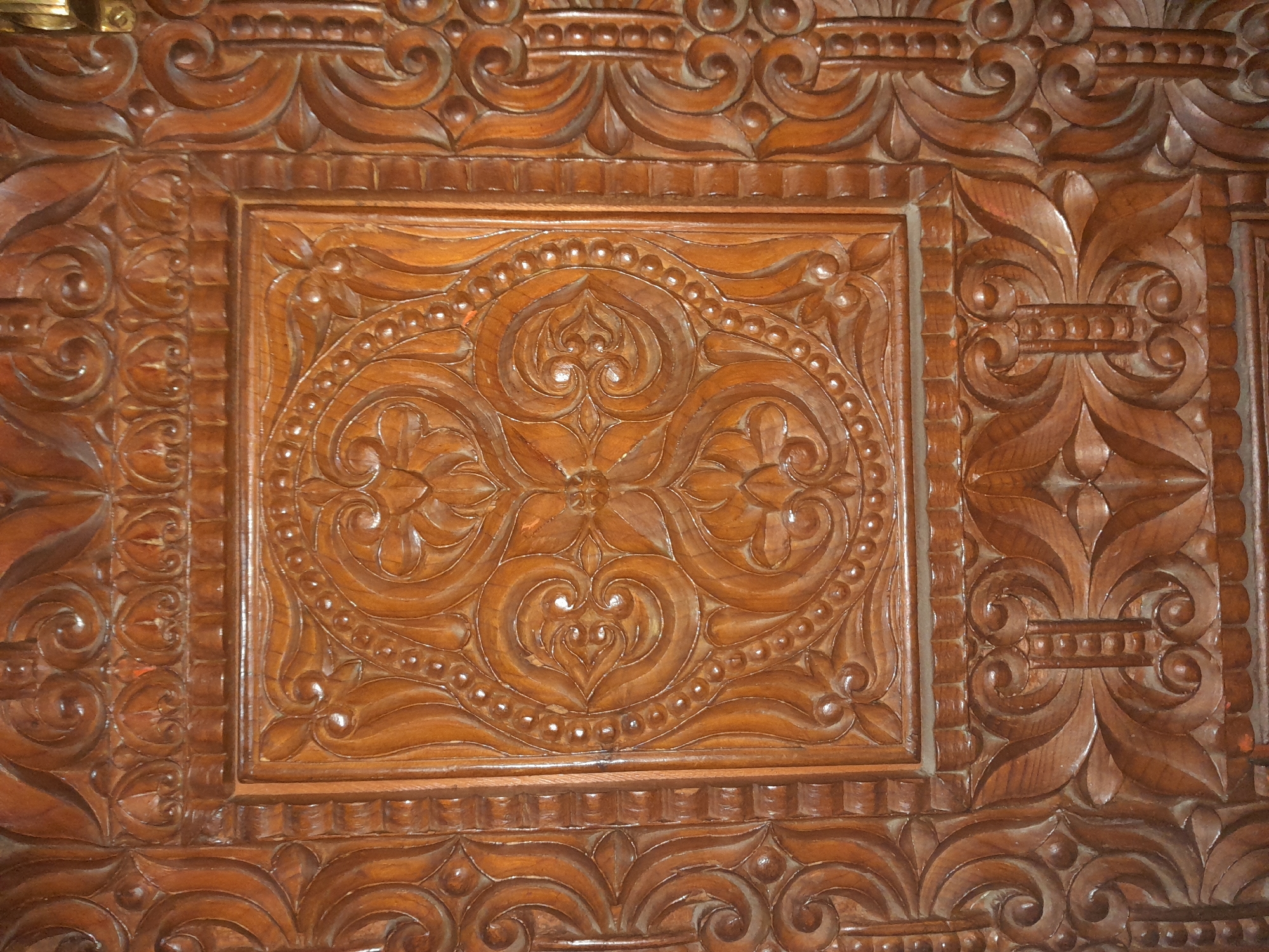 The image shows a close-up of intricately carved dark wood. The carving features geometric patterns and floral motifs, with a central square panel showcasing a symmetrical floral design surrounded by a beaded border. The wood appears to be a door or a piece of furniture. The lighting highlights the depth and texture of the carvings. There are no people or identifiable landmarks in the image.