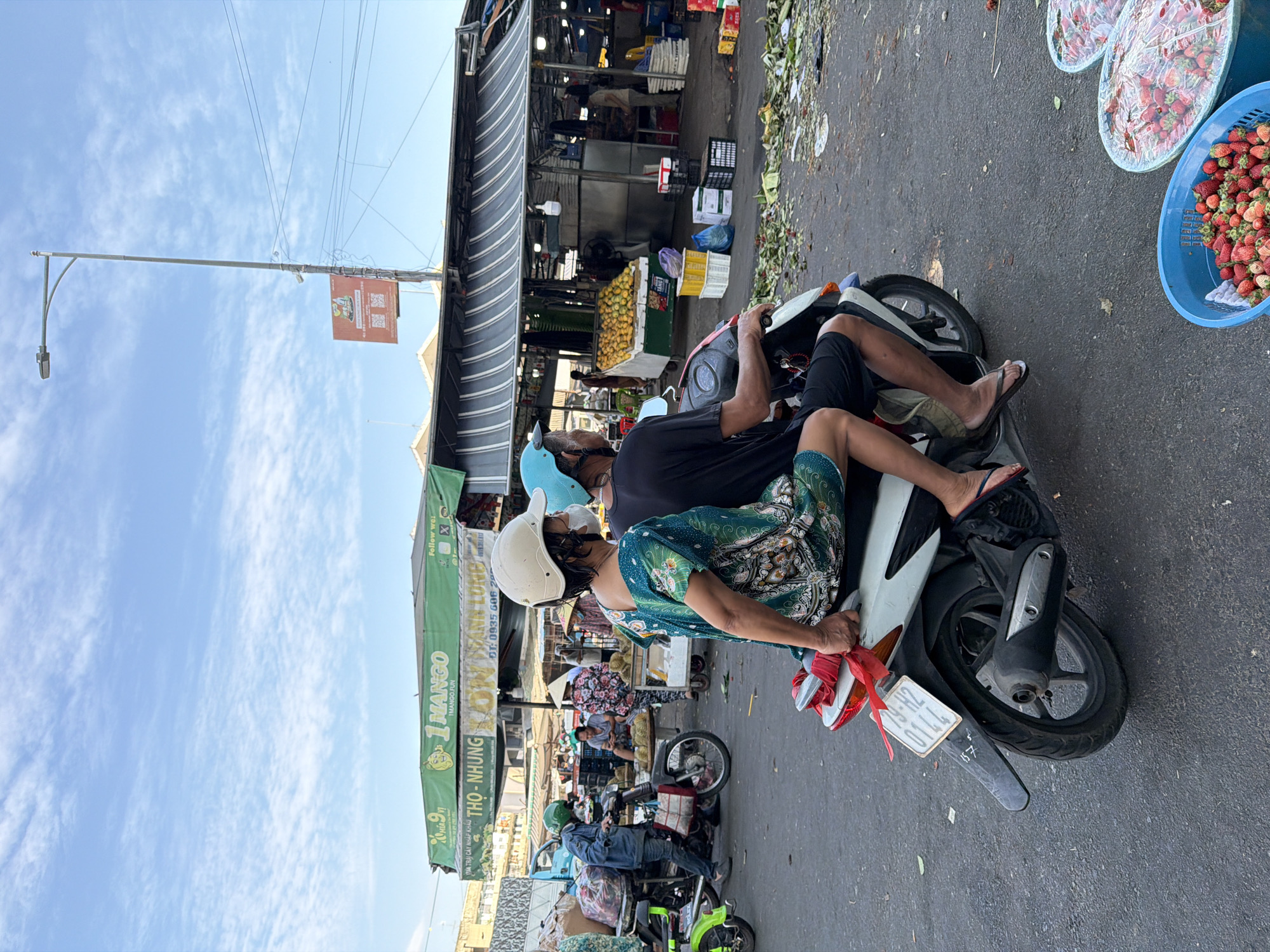 The image captures a street scene in Nha Trang, Vietnam, likely at a market. Two individuals, a woman in a green patterned dress and a man in a black t-shirt, are seated on a white scooter. The woman, in the foreground, is wearing a white helmet and a face mask, with her right hand holding onto the back of the scooter. The man is also wearing a helmet and appears to be operating the scooter.
The environment is characterized by a marketplace setting. A green awning displaying "1 MANGO" is visible in the background, suggesting a business or stall. Various market stalls with awnings are present, some displaying produce, including a large pile of yellow fruits, likely mangoes, and baskets filled with strawberries. Several people are scattered throughout the market, some interacting with vendors and others moving around. The ground is paved and shows signs of activity, with fallen leaves and debris. A street light is visible in the upper right portion of the image, and overhead wires are present. The sky is a clear blue with scattered white clouds. A license plate on the scooter reads "79-H2 0144".