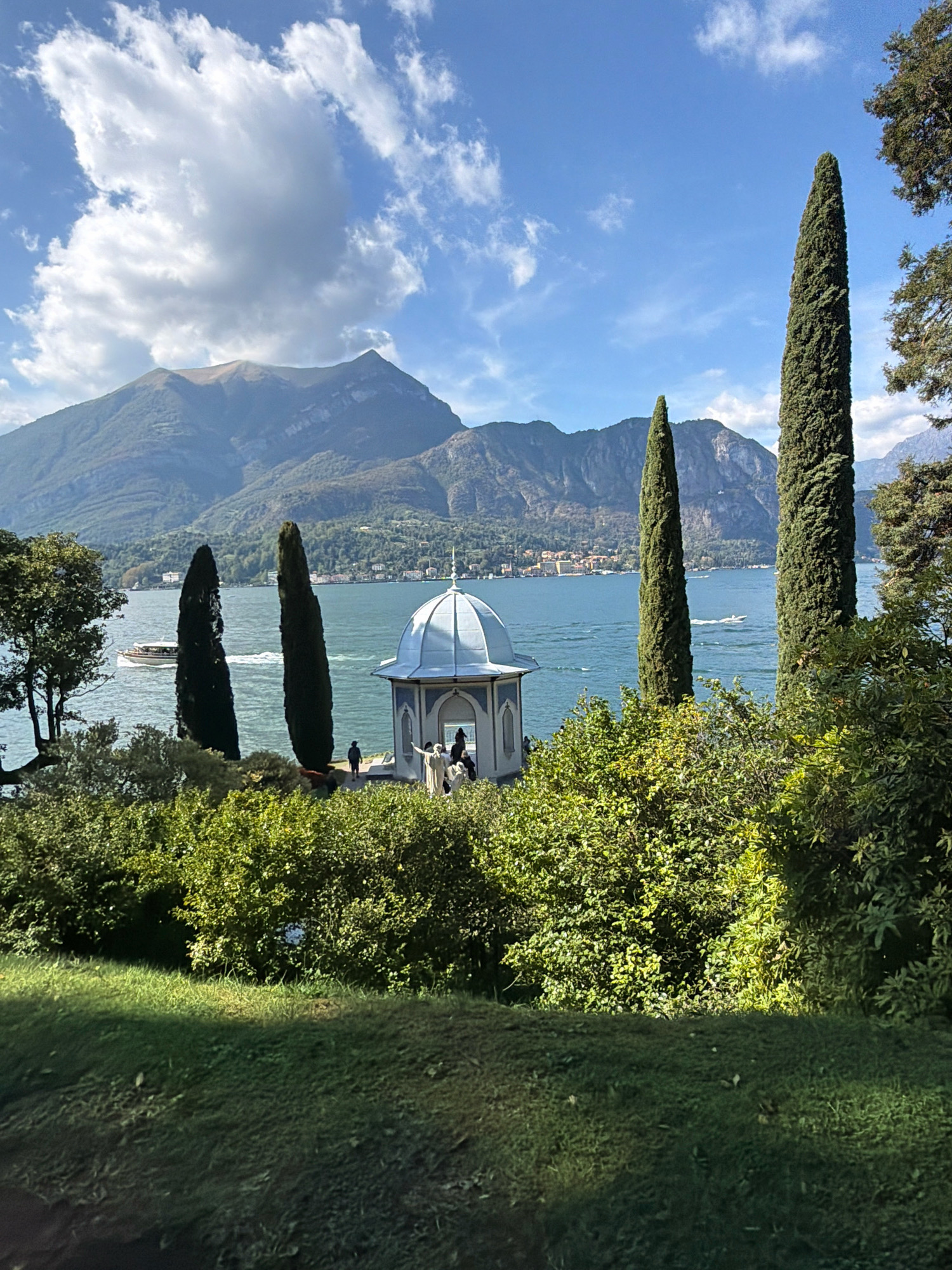 A scenic view of a lake and mountains is captured on a sunny day. In the foreground, lush green bushes and a manicured lawn occupy the lower portion of the frame. The middle ground features a tranquil body of water, a pavilion with a domed roof, and several tall, slender cypress trees. A boat is visible on the lake, moving away from the viewer. In the background, majestic mountains rise, their peaks partially obscured by fluffy white clouds against a clear blue sky. The scene evokes a sense of peacefulness and natural beauty, consistent with a location such as San Giovanni, Italy, known for its picturesque lakes and surrounding landscapes.