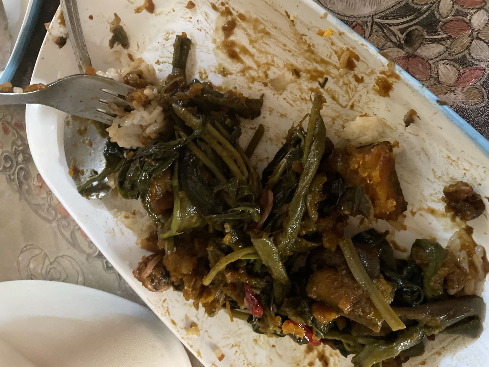 A partially consumed meal is depicted on a white rectangular ceramic plate, indicating a dining setting in Davao City, Philippines. The plate contains a mix of green leafy vegetables, identifiable as kangkong (water spinach), and chunks of orange-brown squash or sweet potato, all coated in a dark, glossy, savory sauce. Small pieces of red chili are interspersed within the dish. White, short-grain rice is mixed with the other components, particularly visible around a metallic fork that is plunged into the food in the upper central portion of the plate. A metallic spoon is partially visible beneath the food on the upper left side. The plate's surface exhibits smears and residue from the sauce and food, indicating active consumption. To the left, a segment of a white, curved ceramic or plastic container is visible. Beneath the plate, a patterned tablecloth or placemat with floral or abstract designs in shades of brown, pink, and light blue provides a textural contrast. A slim, blue-edged object, possibly another plate or tray, is partially visible in the upper right background. No individuals are present in the image.