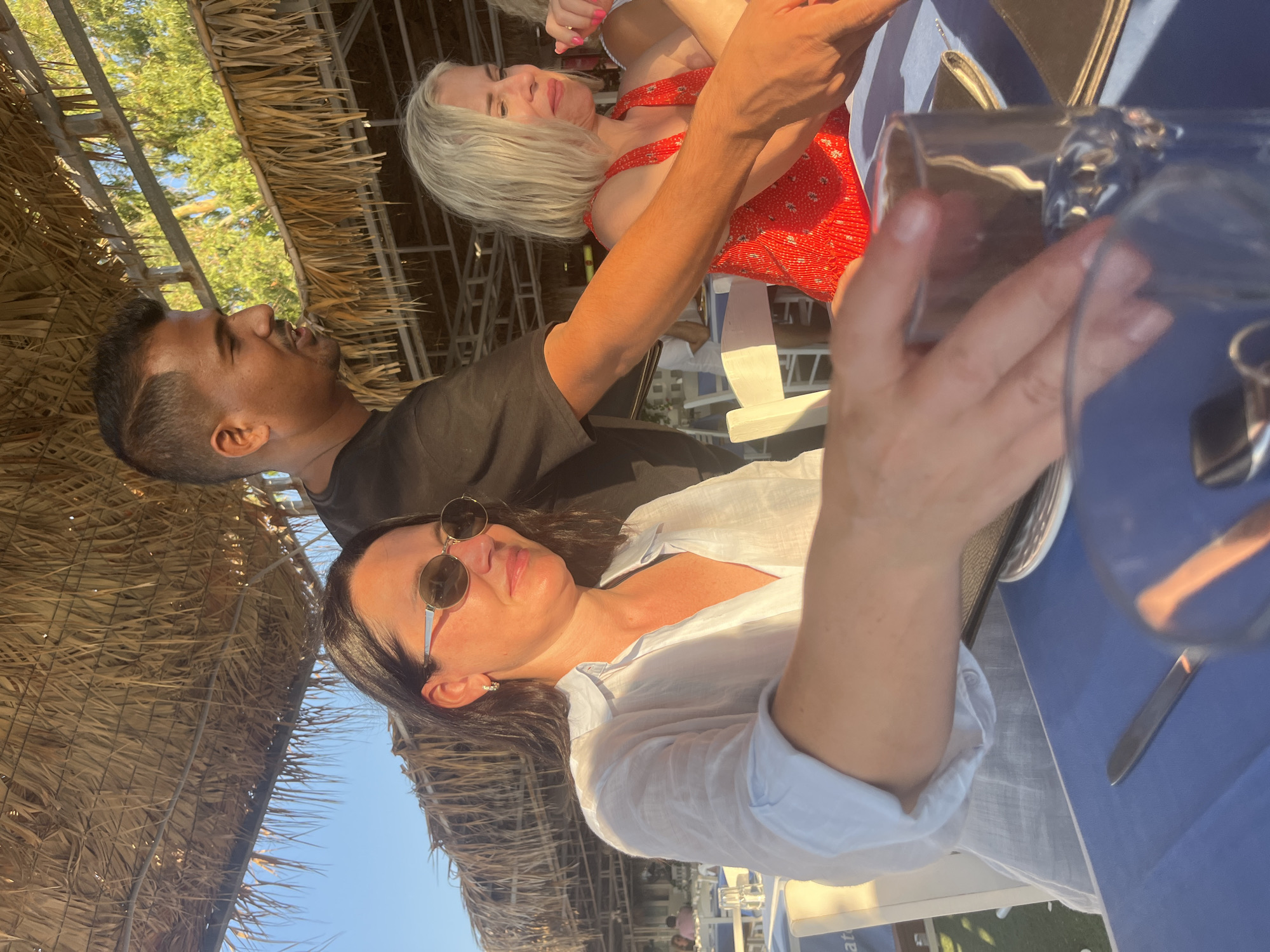 A group of people are dining outdoors under a thatched roof. A man in a black t-shirt is serving drinks to two women. The woman on the left is wearing a white linen shirt and sunglasses, and the woman on the right is wearing a red dress. They are seated at a table with blue tablecloths. The scene appears to be a restaurant in Polis Chrysochous, Cyprus, given the presence of Mediterranean vegetation and the overall ambiance.