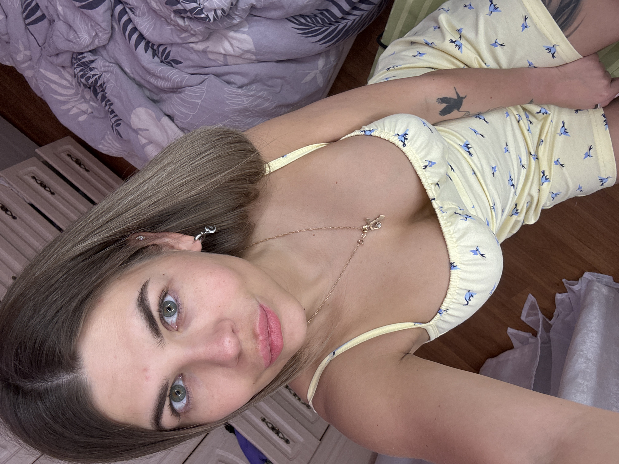 A woman with light brown hair and green eyes is lying down in a room. She is wearing a yellow dress with blue bird patterns and a gold necklace. There is a bird tattoo on her left arm. The room contains a bed with a gray and white patterned duvet, a radiator, and white drawers. The location is Chaikovsky, Russia.