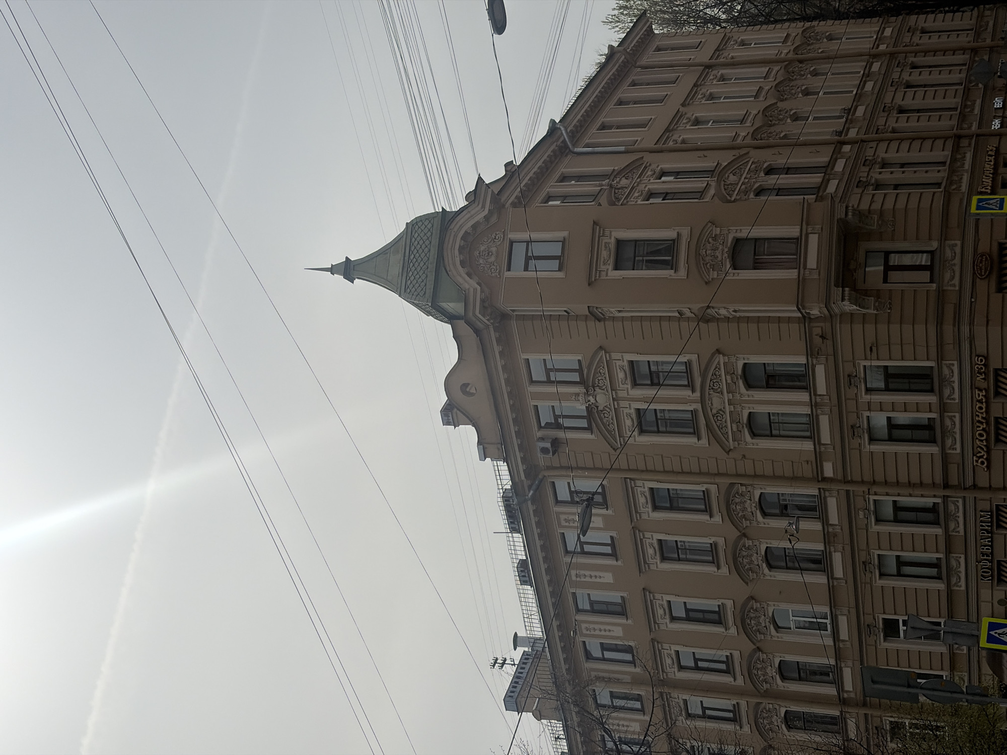A low-angle view showcases the corner of a multi-story building in Saint Petersburg, Russia. The building features a beige façade, ornately decorated with architectural details around the windows. A small, green-roofed turret tops the corner of the building. Visible on the lower right of the building is Russian text; a pedestrian crosswalk sign is also present. Above, a network of power lines stretches across the sky, which is hazy and bright with the sun. The angle of the image is tilted slightly.