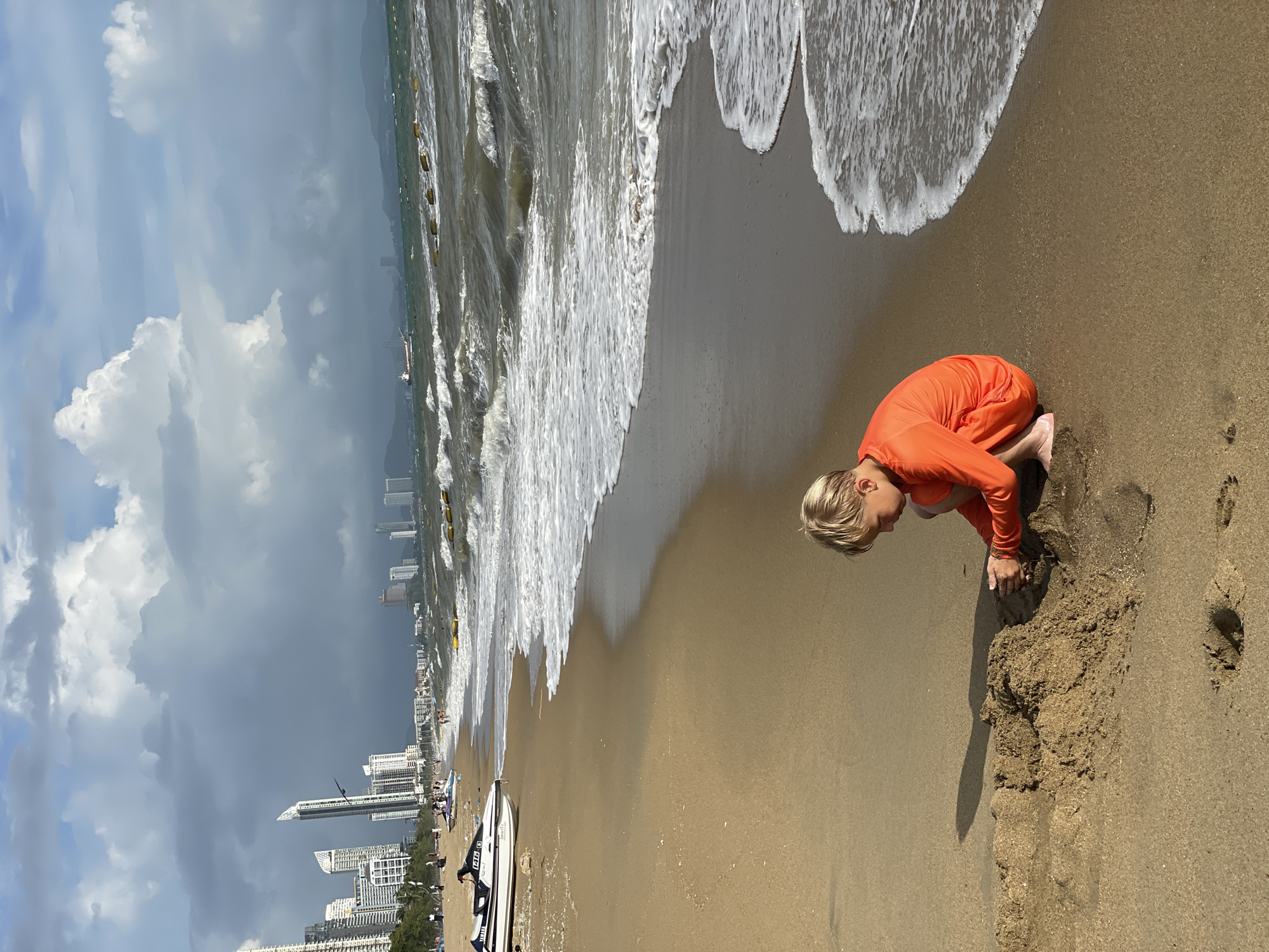 A young boy with blonde hair, wearing an orange long-sleeved shirt and dark shorts, is playing in the sand on a beach in Pattaya City, Thailand. He is crouching and digging in a sandcastle he is building. The sandcastle is located in the foreground. The ocean with waves is to his right, and the beach extends to the left, where several jet skis are parked. In the background, there are tall buildings that are characteristic of Pattaya City, and the sky is partly cloudy with dramatic cloud formations.