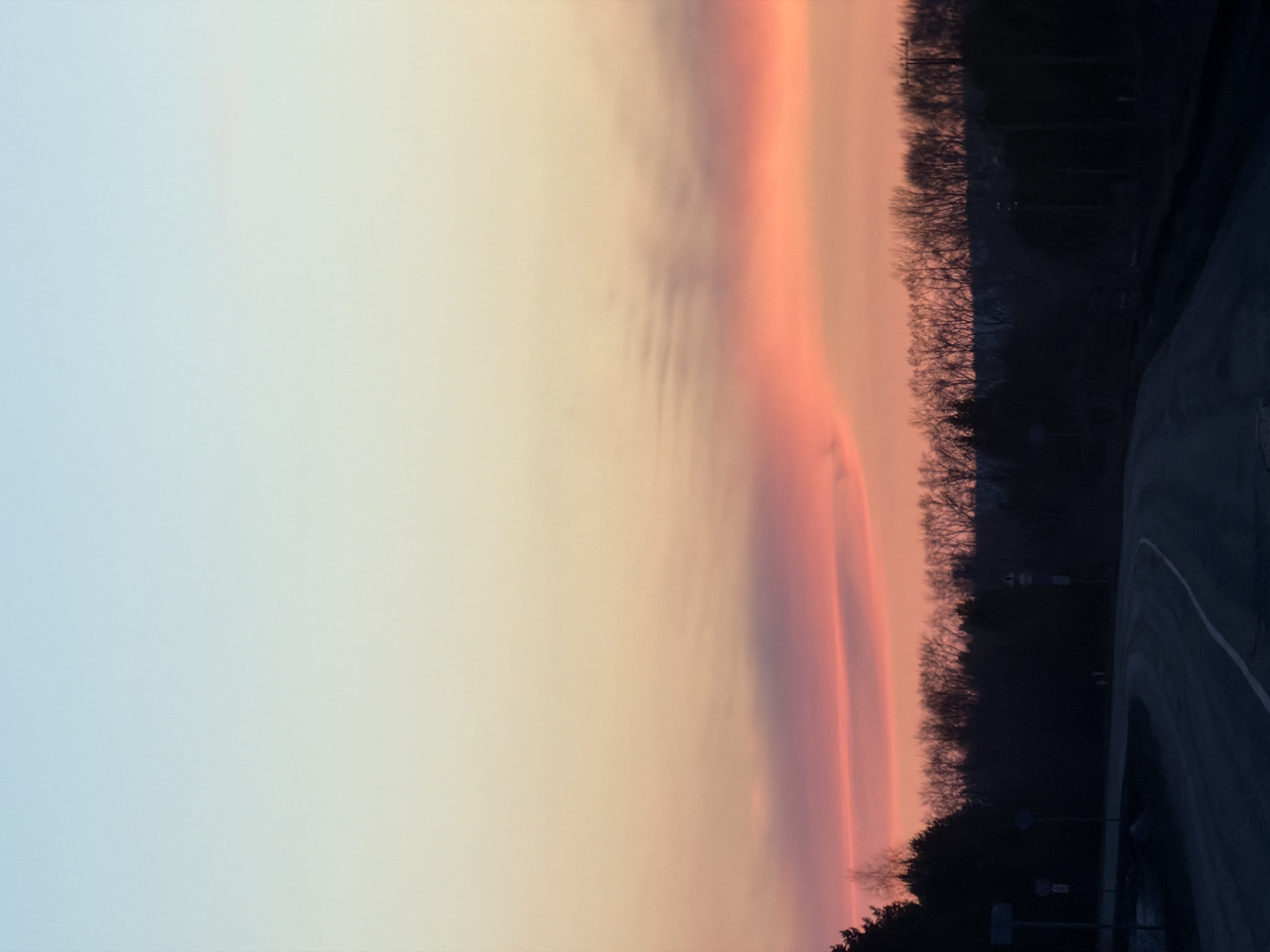 The image shows a sunset sky in Gory, Russia. The sky transitions from light blue on the left to orange and pink near the center, where a thick band of reddish-purple clouds is present. To the right, a dark treeline forms the edge of a forest or park. A section of road with a white dividing line is visible in the lower right corner.