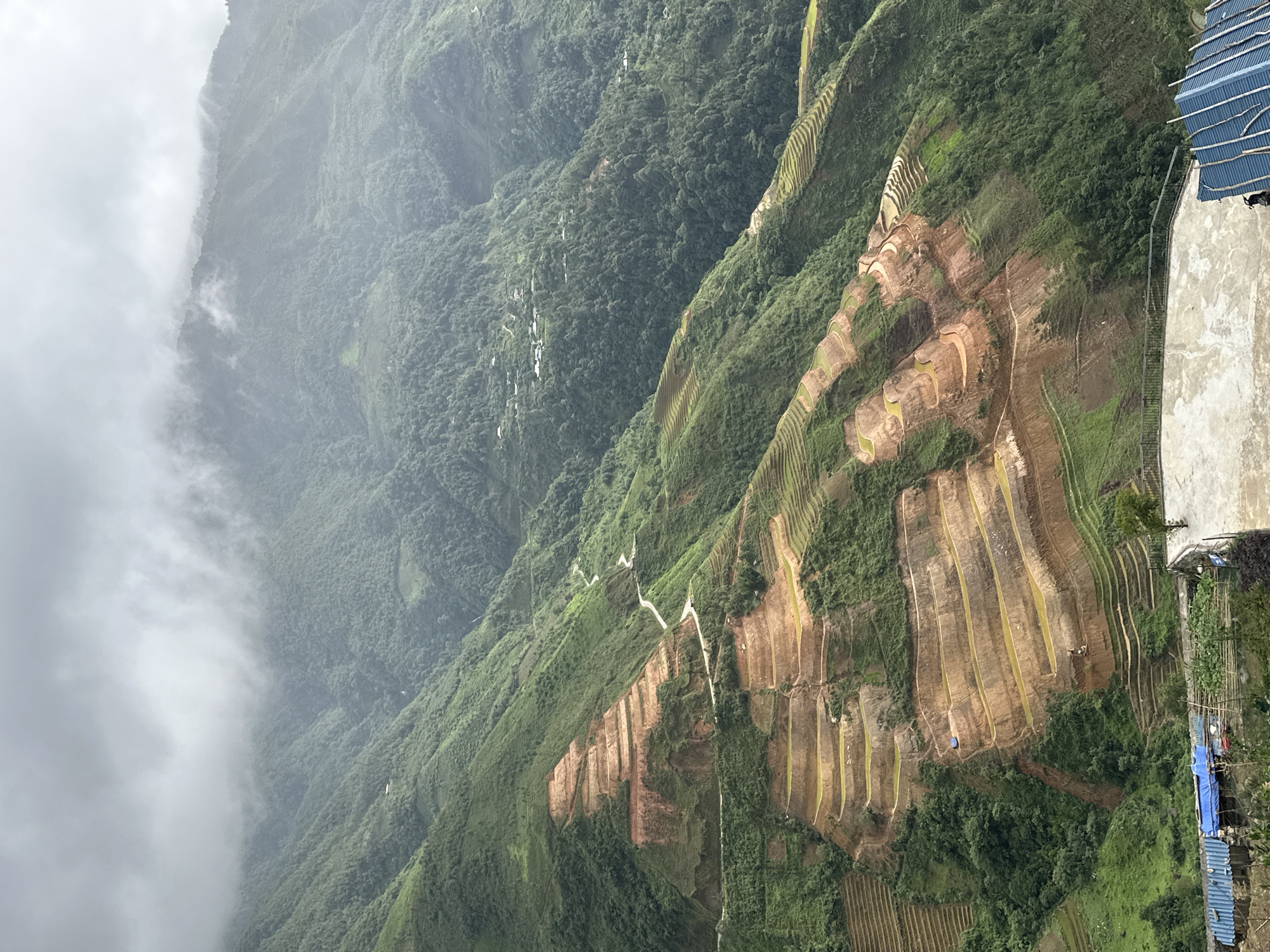 The image shows a panoramic view of terraced rice paddies on a steep mountainside, characteristic of the landscape in Ta Xua, Vietnam. The mountains are covered in lush green vegetation, with some areas of exposed reddish-brown soil revealing the terracing. A winding dirt road snakes its way through the terraces, connecting different parts of the landscape. The sky is overcast with thick clouds, creating a hazy and diffused light. In the foreground, a concrete platform with a metal railing overlooks the valley. Several small structures with blue corrugated roofs are visible at the bottom of the frame, suggesting human habitation or agricultural activity. The overall scene depicts a rural, mountainous environment with extensive agricultural development.