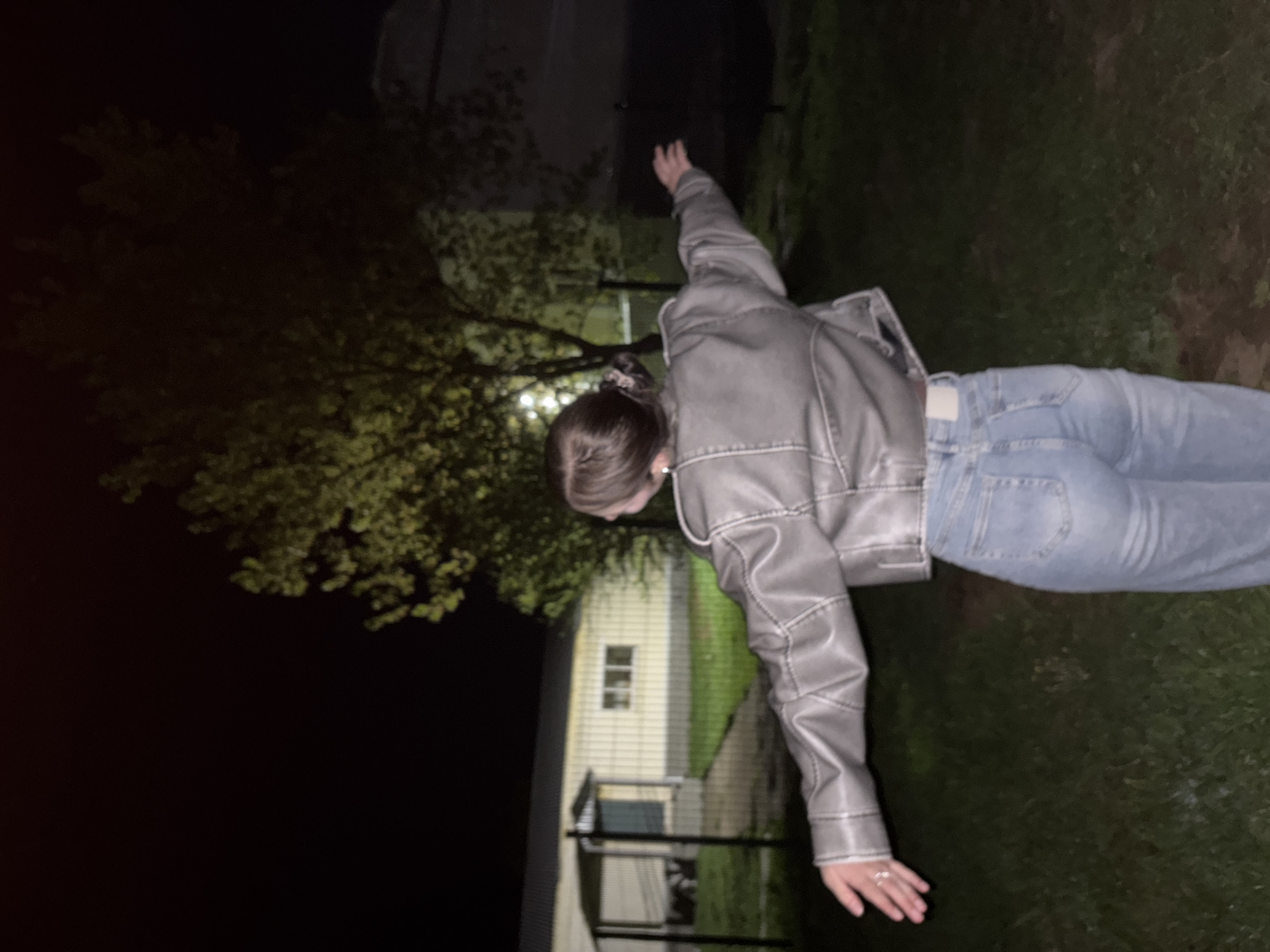 A full-body shot of a person in a light gray jacket and blue jeans is standing outside on a grassy area at night. There's a tree to the left with a building behind it. The location is Фоки, Russia, however I can not confirm due to lack of evidence.