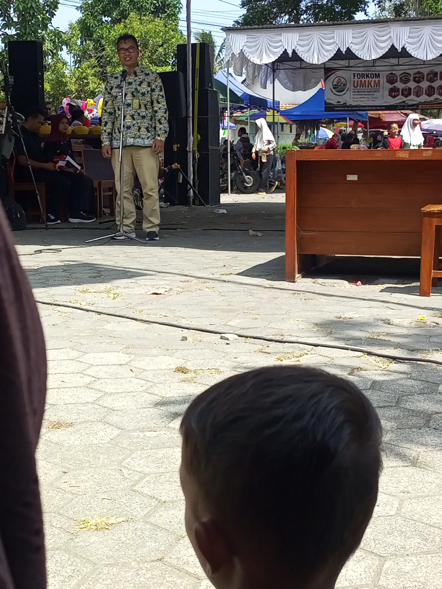 The image captures an outdoor event in Nanggulan, Indonesia. A man in a patterned shirt and khaki pants stands at a microphone on a stage, addressing an unseen audience. To his left, two individuals are seated, one in black and the other in a red headscarf. Behind the stage, tents and stalls suggest a market or community gathering. The ground is paved with hexagonal tiles. In the foreground, the back of a child's head is visible, suggesting they are observing the event from a lower vantage point. The environment appears sunny with trees in the background. A banner on the stage reads "FORKOM UMKM KALIMAHAN BANYUROTTO".