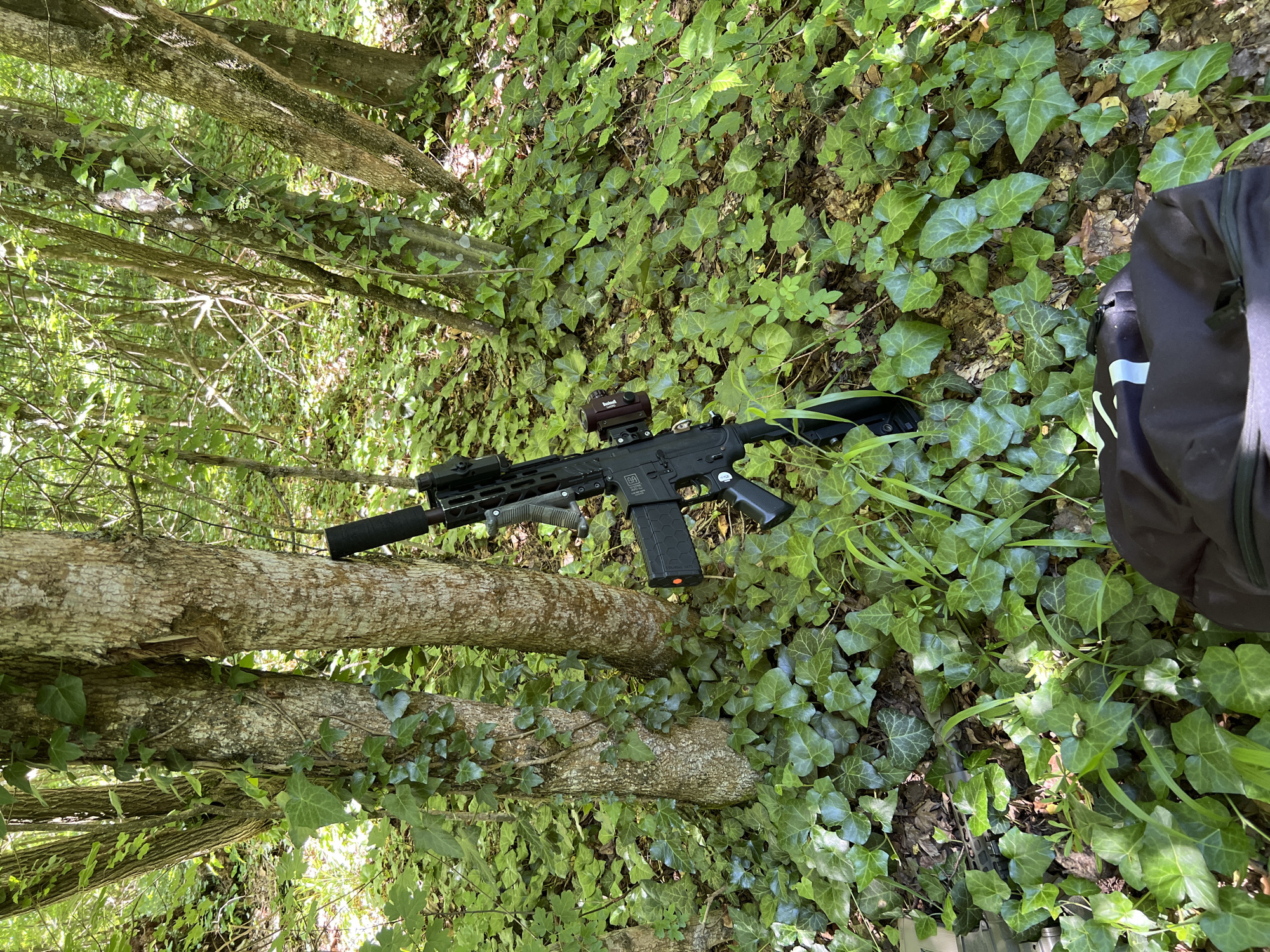 The image depicts a black AR-15 style rifle lying on a fallen tree trunk amidst dense green foliage, possibly ivy. Part of a black backpack is visible to the right of the frame. The location appears to be a wooded area, but the city and specific location are unknown.