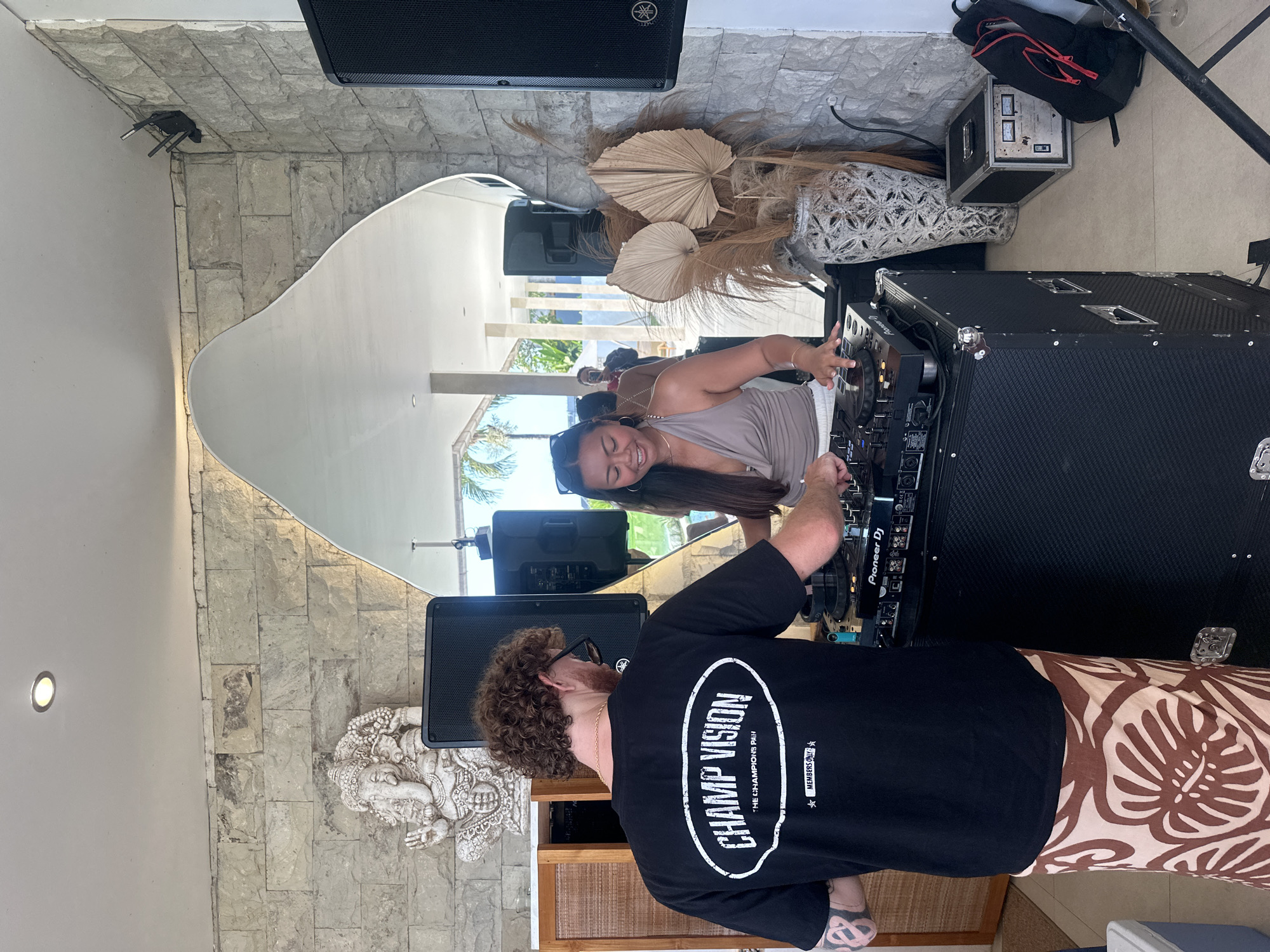 In a semi-open-air venue in Mengwi, Indonesia, two individuals are operating DJ equipment. The primary subject is a male seen from behind, positioned to the left of the frame. He has curly brown hair, wears a black t-shirt with "CHAMP VISION," "THE CHAMPIONS PAIN," and "MEMBERS ONLY" printed in white, brown and cream patterned sarong-style trousers, and black sunglasses resting on his head. Tattoos are visible on his left arm. He stands in front of a black Pioneer DJ mixer/controller unit, which rests on a large black road case.
To the right of the male, a female is also engaged with the DJ equipment, smiling and looking towards the left. She wears a light brown halter top and white bottoms, with black sunglasses pushed up onto her head. Her hands are on the controls of the DJ mixer/controller.
The DJ setup includes two large black Yamaha speakers, one positioned directly behind the male and another to the right, partially obscured by the mirror. An additional silver rack-mount audio unit with three visible meters is on the floor to the right of the main DJ setup, alongside a black backpack with red zipper pulls and a clear glass containing a light-colored liquid.
The background features a textured stone wall composed of light-colored, rectangular blocks. On the left, a white statue of Ganesha is visible on a wooden cabinet. Above the Ganesha statue, a recessed ceiling light is illuminated. A large, irregularly shaped mirror occupies the center-right of the wall, reflecting an outdoor area with pillars, palm trees, and distant figures. To the right of the DJ setup, a decorative arrangement of dried palm fronds and other dried foliage is placed in a textured white vase on the tiled floor. The floor is light-colored tiled, and a white cooler is visible on the far left.