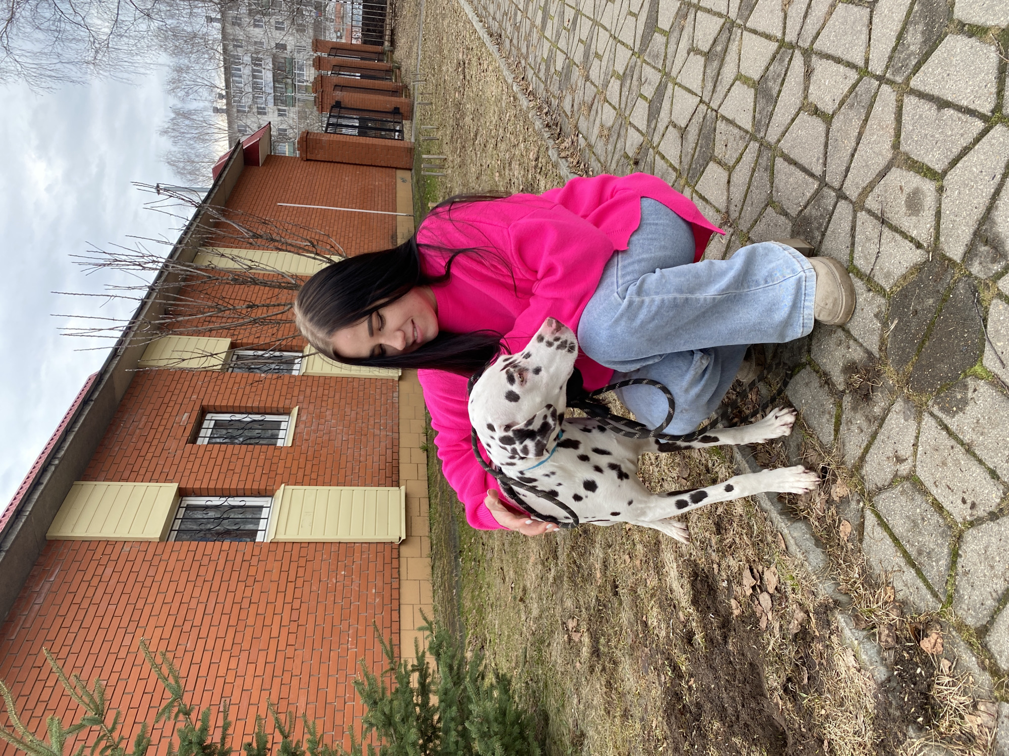 A young woman in a bright pink sweater and light blue jeans crouches on a paved walkway, embracing a Dalmatian dog. The dog is white with black spots and is wearing a blue collar. The woman's hair is long and dark. Behind them is a two-story brick building with a red roof and white-framed windows. The walkway is made of grey paving stones with grass and dead leaves between them. The sky is overcast. Based on the architecture and visual cues, the location is likely Chaikovsky, Russia.