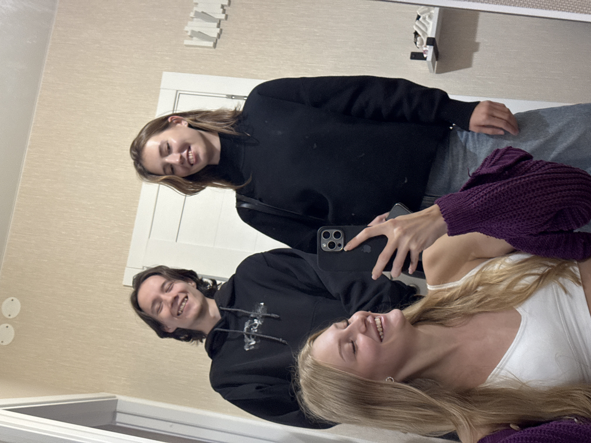 A medium shot captures three young adults posing for a selfie in what appears to be a residential hallway with a cream-colored, textured wallpaper. In the foreground, a young woman with long blonde hair is looking at the camera, smiling widely. She is wearing a white tank top and a purple knitted cardigan draped over her shoulders. She holds a smartphone in her right hand, which is capturing the photo. Behind her and slightly to her left, a young man with dark, shoulder-length hair is smiling and looking at the camera. He is wearing a black hooded sweatshirt. To his right, a young woman with medium-length brown hair and a black turtleneck sweater is also looking at the camera and smiling. She is wearing a black crossbody bag over her left shoulder. The background shows a white door frame and a white closet or door. On the right side of the frame, a white shelf is mounted on the wall, displaying several items, possibly toiletries or decorative objects. Two round, white fixtures are visible on the wall above the man's head. The lighting is diffused and appears to be artificial. The overall atmosphere is casual and friendly. The image appears to have been taken inside a residential building in the city of Perm, Russia, given the context.