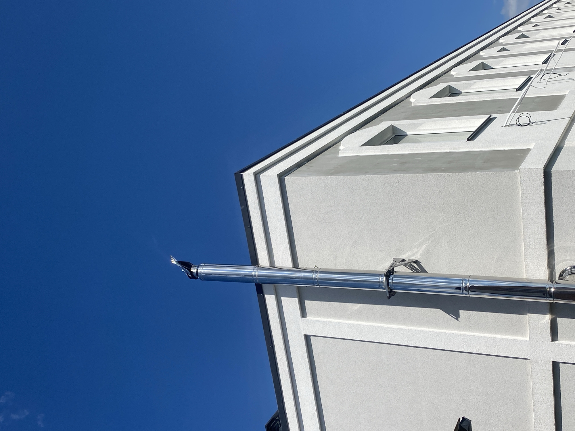 The image shows a modern building with a white exterior and dark trim, possibly in Novi Petrivtsi, Ukraine. A tall, reflective metal chimney extends vertically upwards along the side of the building, against a clear blue sky. The building features several rectangular windows with dark frames. The overall scene suggests a new construction or renovation, with a focus on clean lines and contemporary design. There are no people visible in the image.
