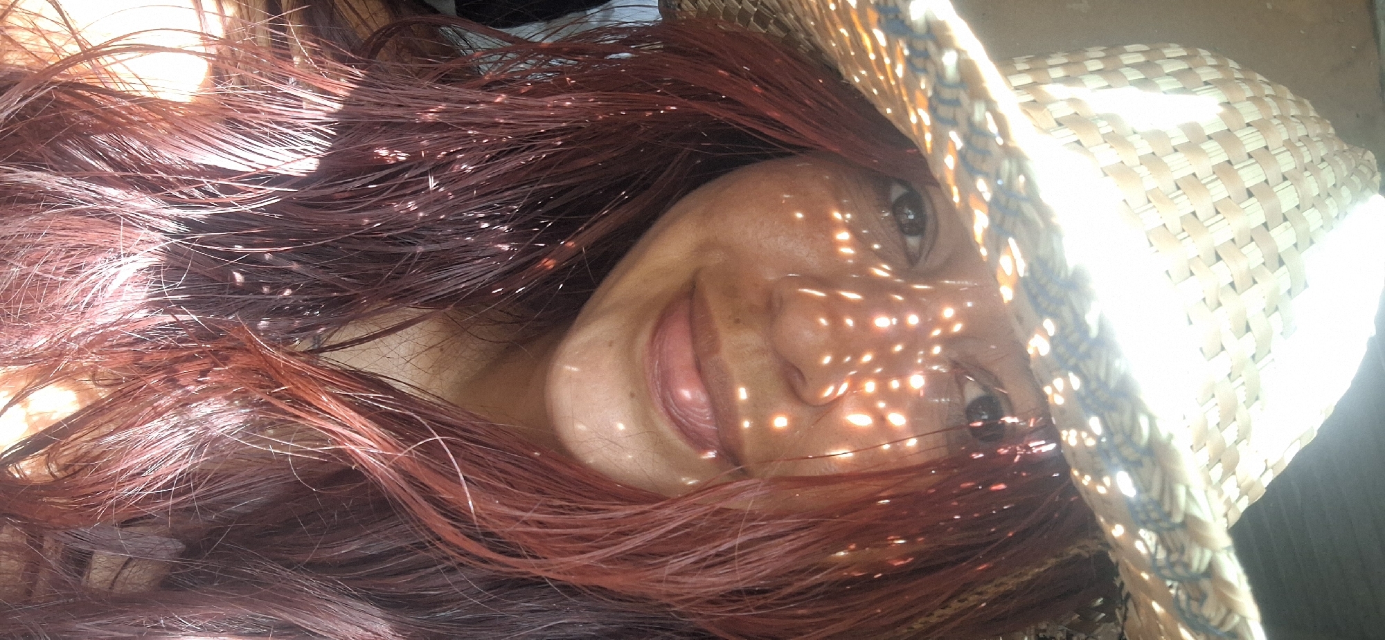 A woman lies down, facing the camera, wearing a straw hat with a woven pattern that casts dappled light across her face. Her hair is reddish-brown and surrounds her head. The scene is brightly lit. There is no explicit indication of the location being in Vallecito, Venezuela, or any elements that would make it identifiable as such.