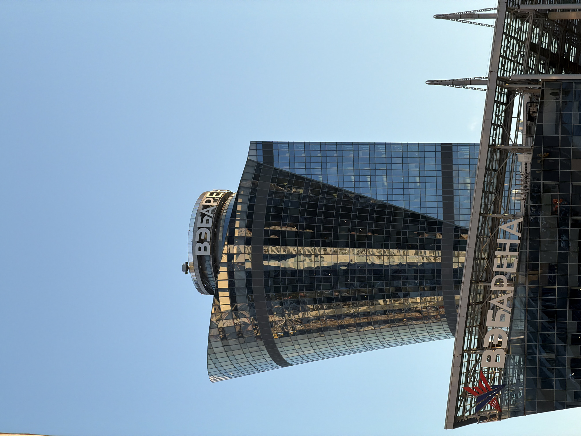 This is a close-up, eye-level, outdoor, daytime, medium shot of a modern, glass-fronted skyscraper with a curved and angled design. The building is predominantly gray and blue, with reflections of the sky and possibly other buildings visible on its glass facade. The top of the building features a circular structure with text that reads "ВЭБ.РФ". Below the main tower, a lower section of the building has a similar glass facade and a metal structure extending outwards, also bearing the text "ВЭБ.РФ" and a red and blue logo. The sky above is clear and blue, with no clouds visible. The scene appears to be in an urban environment, but the specific city is not discernible from the image.