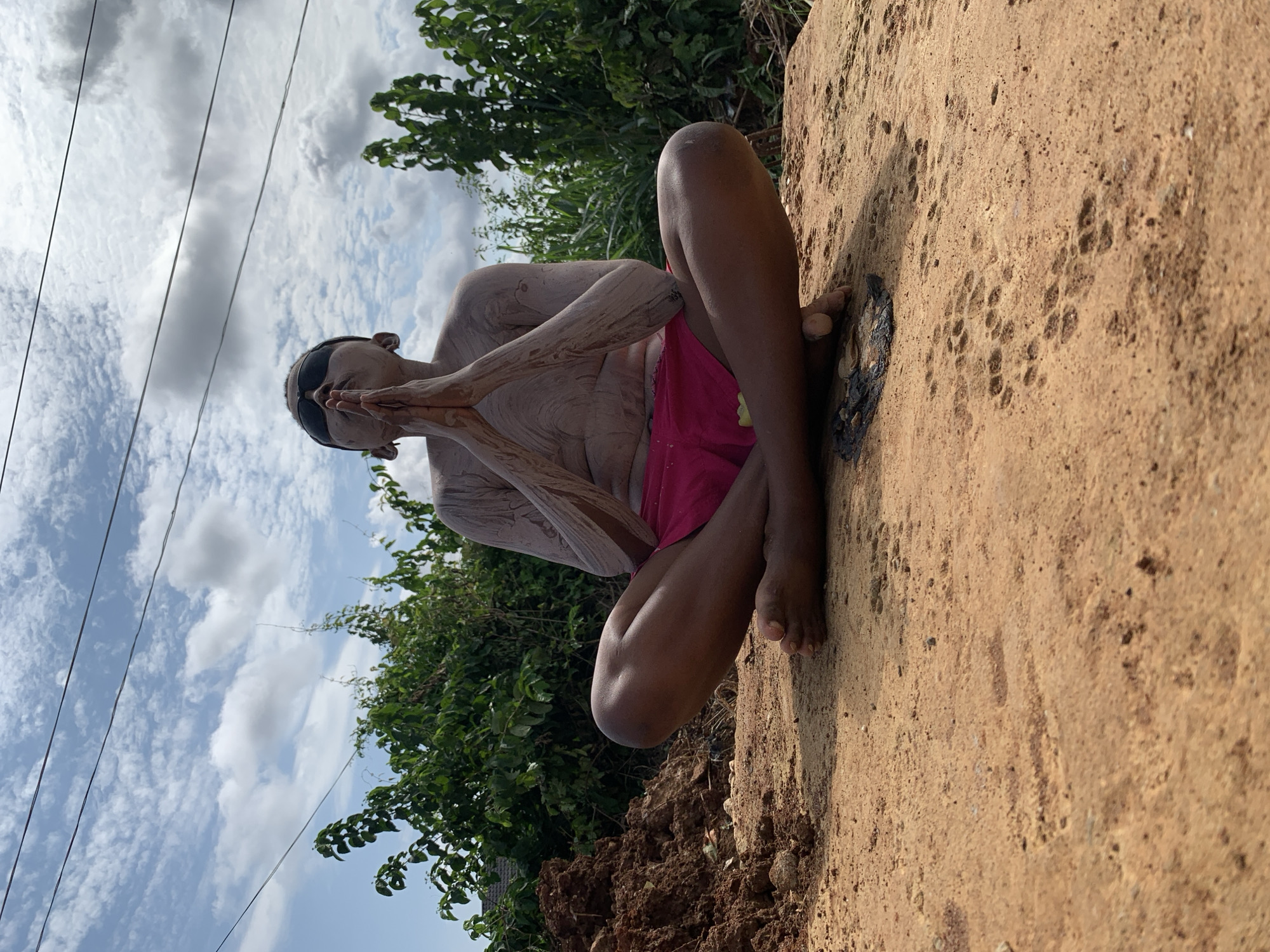 A person is seated in a meditative pose on a large, flat, light brown rock. They are wearing dark sunglasses and pink shorts. Their body is covered in a light-colored substance, possibly mud or ash, with streaks and patterns visible on their arms, torso, and legs. Their hands are pressed together in front of their chest in a prayer-like gesture.
The scene is outdoors, with lush green foliage and trees visible in the background. The sky is a bright blue with scattered white clouds. Power lines are visible stretching across the sky. The ground appears to be dirt, with some reddish-brown soil and rocks. The lighting suggests it is daytime, likely late morning or early afternoon given the brightness of the sun and the angle of the shadows. The overall mood is serene and contemplative.
The location context provided is Ikorodu, Nigeria. While not explicitly visible in the image, the presence of lush vegetation and the general atmosphere are consistent with a tropical or sub-tropical environment. No visible text is present in the image.