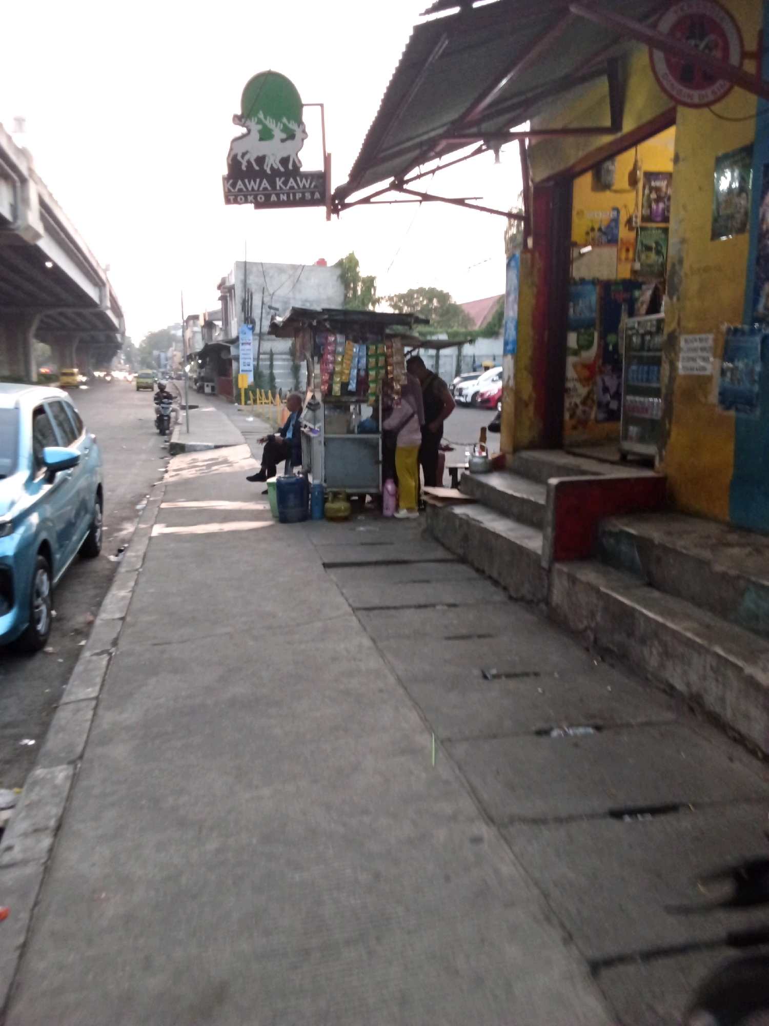The image depicts a street scene in Bandung City, Indonesia, likely in a commercial or mixed-use area. A bright blue car is parked on the left side of the road, facing away from the camera. To the right of the car, a concrete sidewalk runs alongside the road.
Along the sidewalk, there is a small food stall or kiosk with a green awning. Two individuals are interacting at the stall. One person, wearing a light-colored top and yellow pants, is standing and facing the stall. Another person, dressed in a dark top and pants, is seated on a blue container next to the stall. A yellow gas cylinder is also visible near the stall.
Further down the sidewalk, there is a multi-story building with a yellow facade, decorated with posters. Steps lead up to the entrance of this building. A sign with a green logo and the text "KAWA KAWAN TOKO ANIPSA" is visible mounted on a pole, indicating a business. In the background, an elevated road or bridge structure is present, with traffic visible on it. The sky is overcast, suggesting either early morning or late afternoon. The overall impression is that of a bustling urban environment with everyday street life and commercial activity.