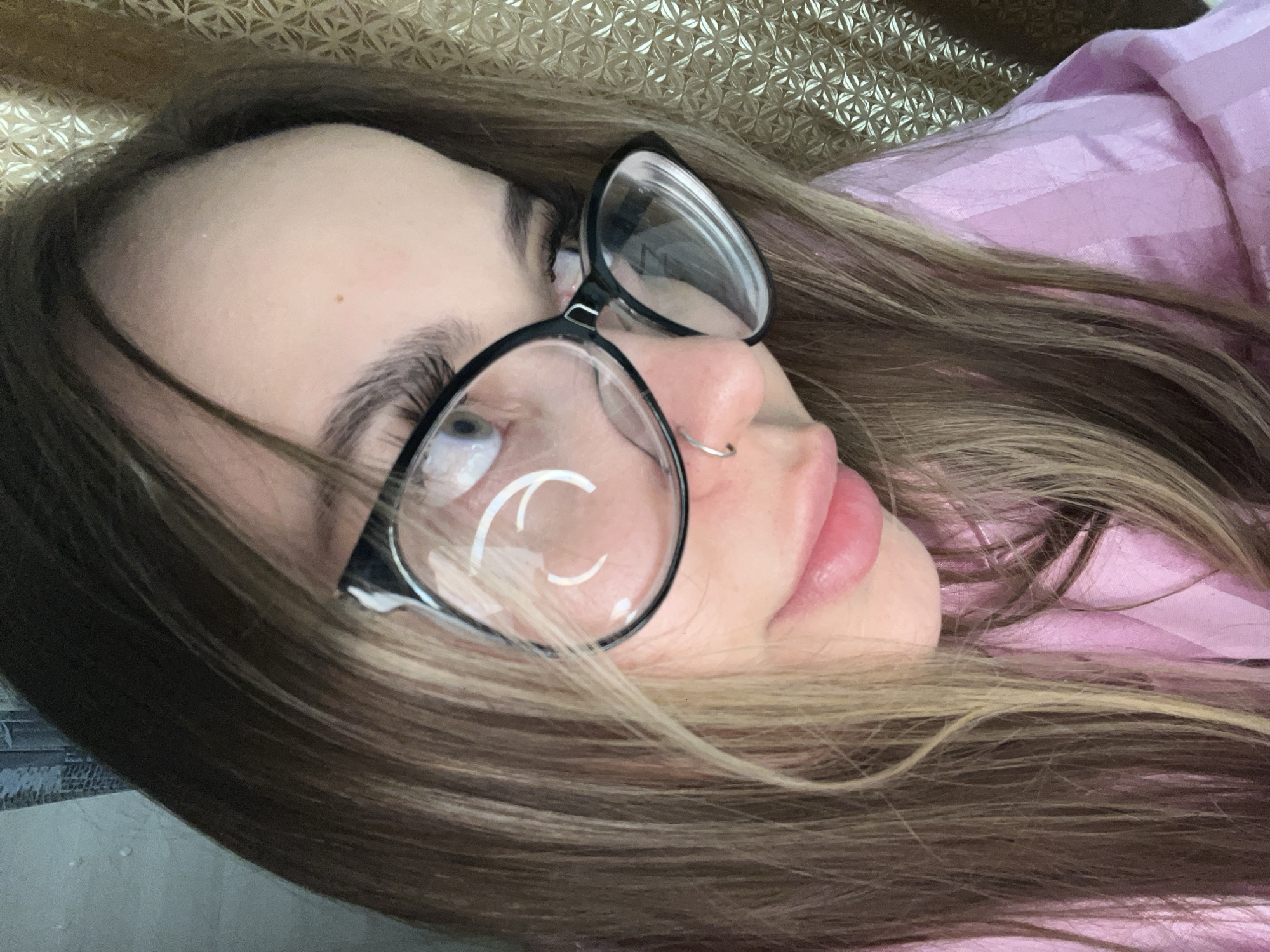 A woman lies with her head tilted back, wearing glasses with black frames and a silver nose ring. She has long, light brown hair and is wearing a light pink shirt. The background is a patterned gold-colored surface. The scene appears to be indoors. There is no indication this photo was taken in Novopokrovskaya, Russia.