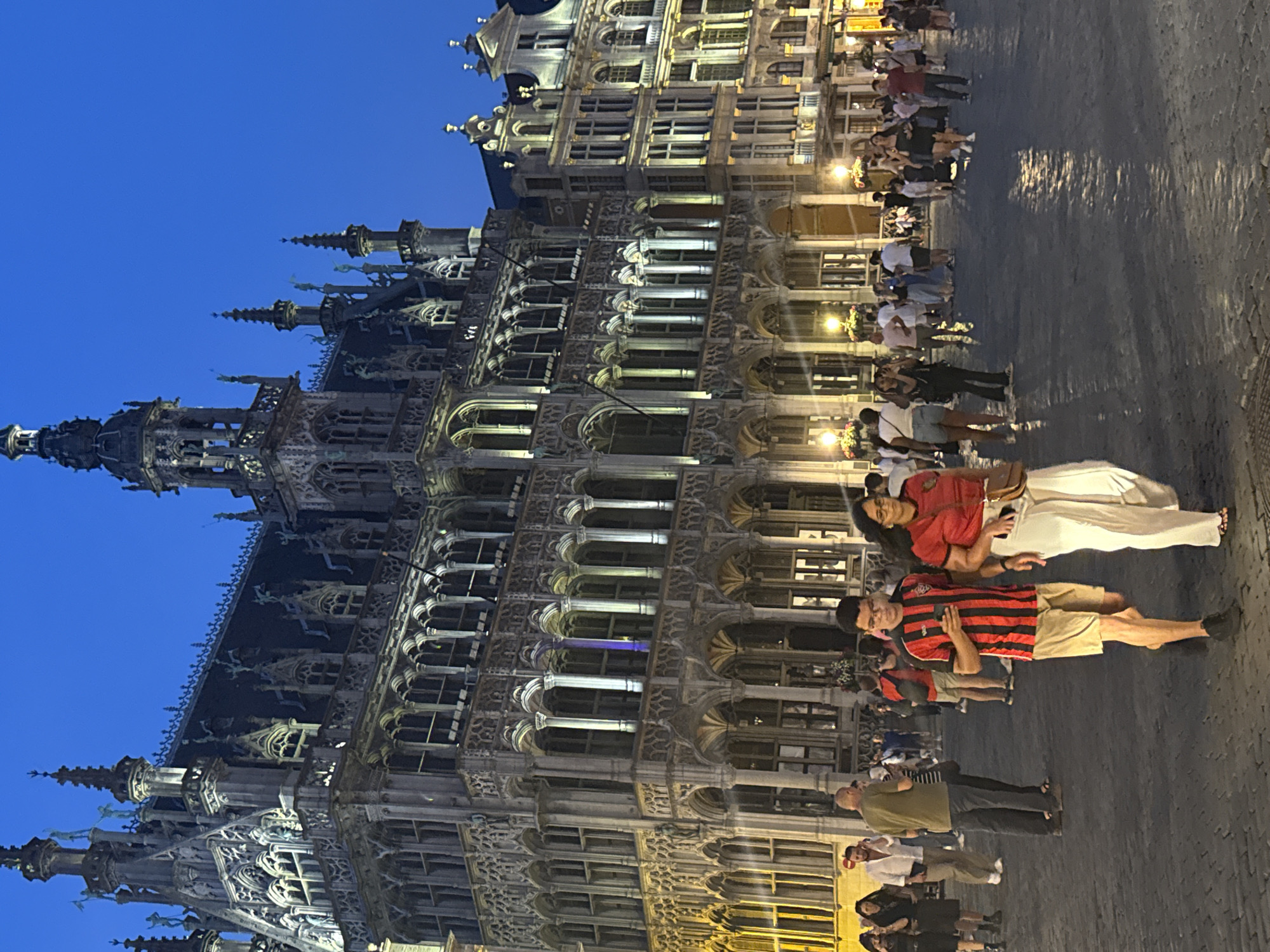 This image depicts a nighttime scene at the Grand Place in Brussels, Belgium. The focal point is the highly ornate Gothic architecture of the Town Hall, illuminated by numerous lights, casting a warm glow on the cobblestone square. The sky is a deep blue, indicating dusk or night.
In the foreground, a young man and a woman are walking towards the camera. The man is wearing a striped red and black t-shirt, tan shorts, and black shoes, and is looking at his phone. The woman is wearing a red t-shirt, white pants, and sandals, and is carrying a brown purse. They are walking on the cobblestone pavement, which reflects the lights from the surrounding buildings.
The square is populated with a moderate number of people, suggesting a lively evening atmosphere. Other individuals are visible in the midground and background, some walking, others standing in groups. The surrounding buildings are also elaborately decorated with architectural details and lights, consistent with historical European city centers. The overall impression is one of a well-preserved historic urban environment at night.