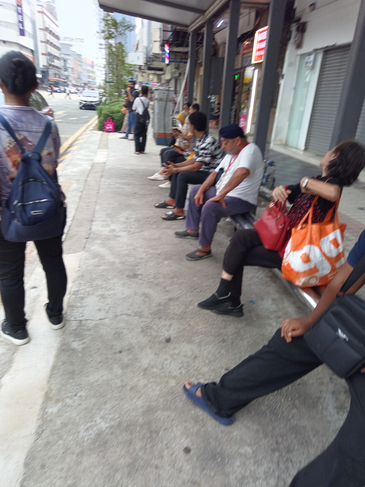 The image depicts a street scene in Kuala Lumpur, Malaysia, likely near a bus stop or a waiting area. In the foreground, a person wearing black pants and black sneakers walks away from the camera, carrying a blue backpack. Several people are seated on a bench in the middle ground, some of whom appear to be waiting. One individual in a black top with red and white patterned accents is looking upwards and holding a red handbag and an orange and white reusable shopping bag. Another person, wearing a white t-shirt and a navy blue headscarf, is seated to their left. Further back, more individuals are visible on the bench, including a person in a black and white patterned shirt and a man in a white t-shirt and a navy blue turban. The background shows a street with traffic, buildings, and a sidewalk. The overall atmosphere suggests a public space in an urban environment, possibly during daylight hours.