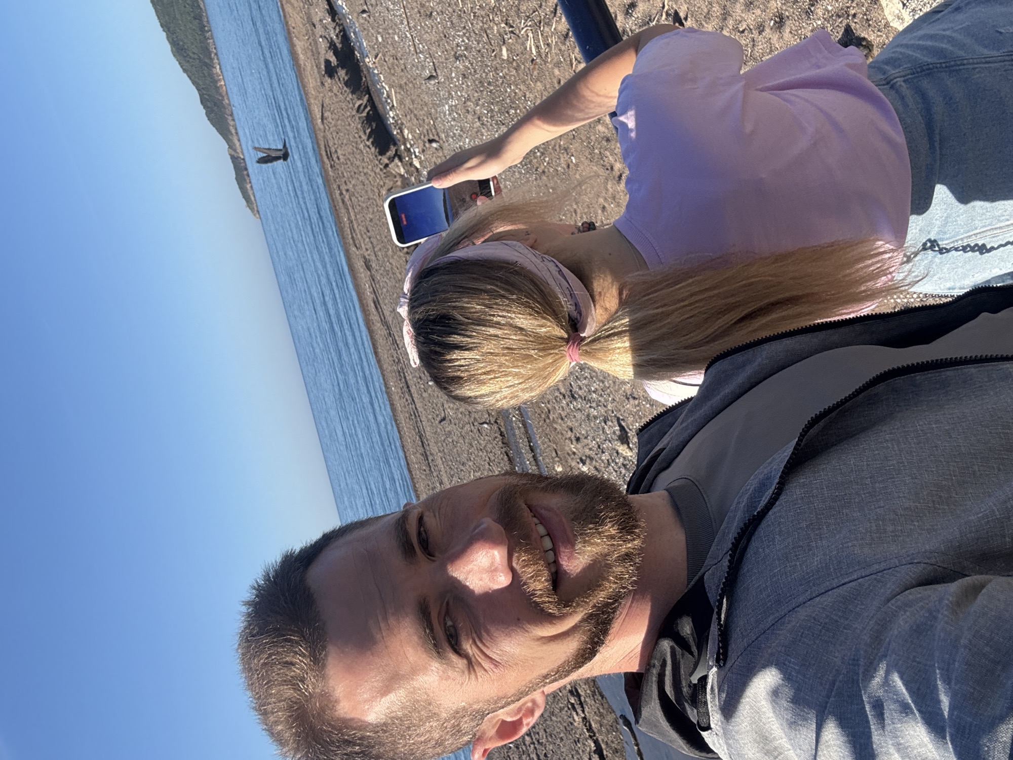A man and a woman are positioned near the coastline, possibly in the vicinity of Boreti, Montenegro, judging by the coastal terrain and water body in the background. The man, with short dark hair and a beard, is closest to the camera and appears to be taking a selfie. He is wearing a grey jacket. The woman behind him has long blonde hair pulled back with a pink band. She is wearing a light purple shirt and is holding a white smartphone, possibly taking a photo of the landscape or the water. The environment includes a pebble or gravel-covered ground, with a coastline and a clear sky visible in the background. In the distance, there is a bird flying above the blue water.
