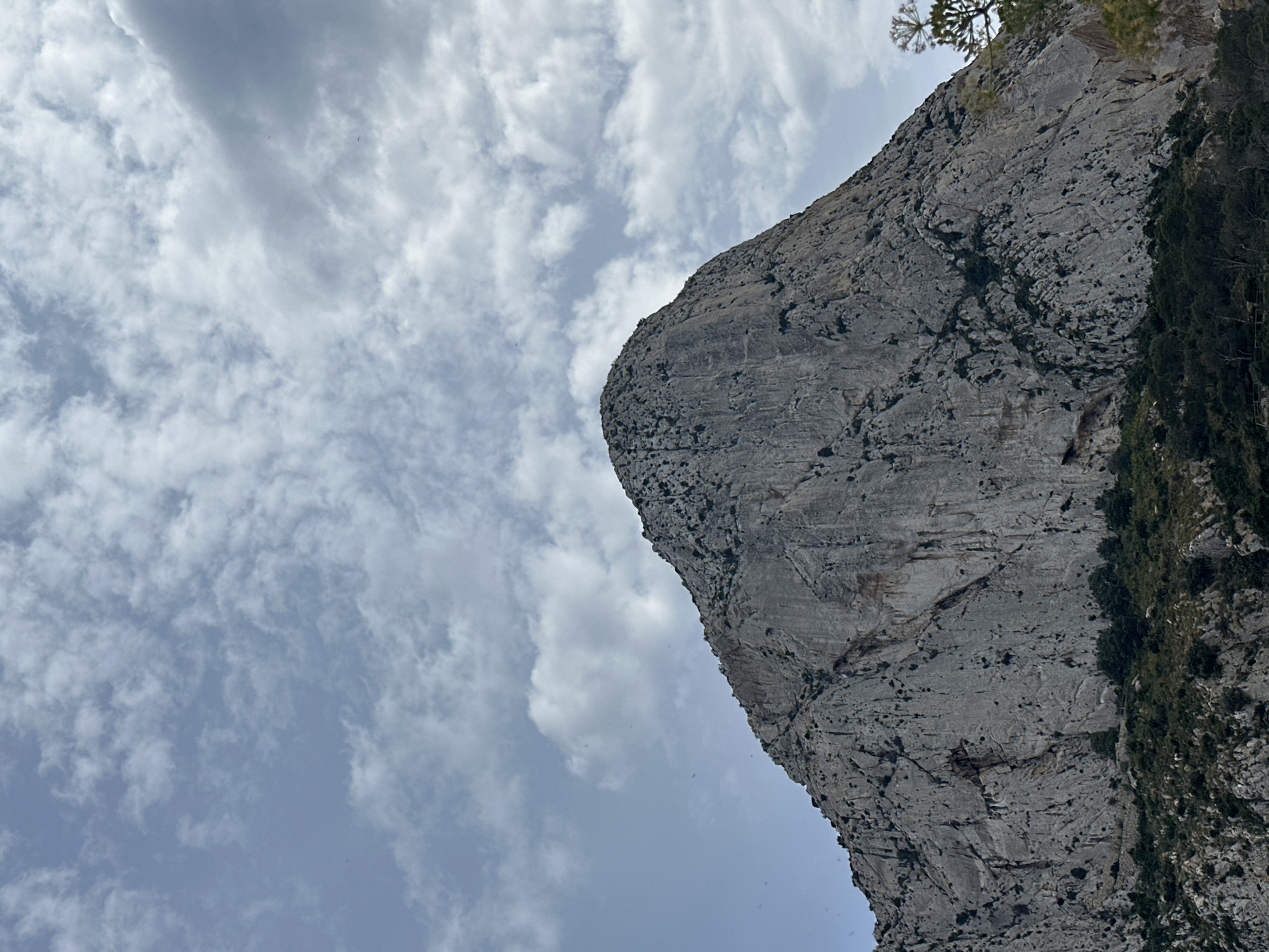 The image shows a gray rock formation extending diagonally from the upper right to the lower right of the frame, positioned against a cloudy sky. The rock surface is textured with crevices and some vegetation growth on the right side. The sky has scattered white and gray clouds against a light blue background. The location in San Gabriel, Spain, is inferred but not visually confirmed. There are no people visible.