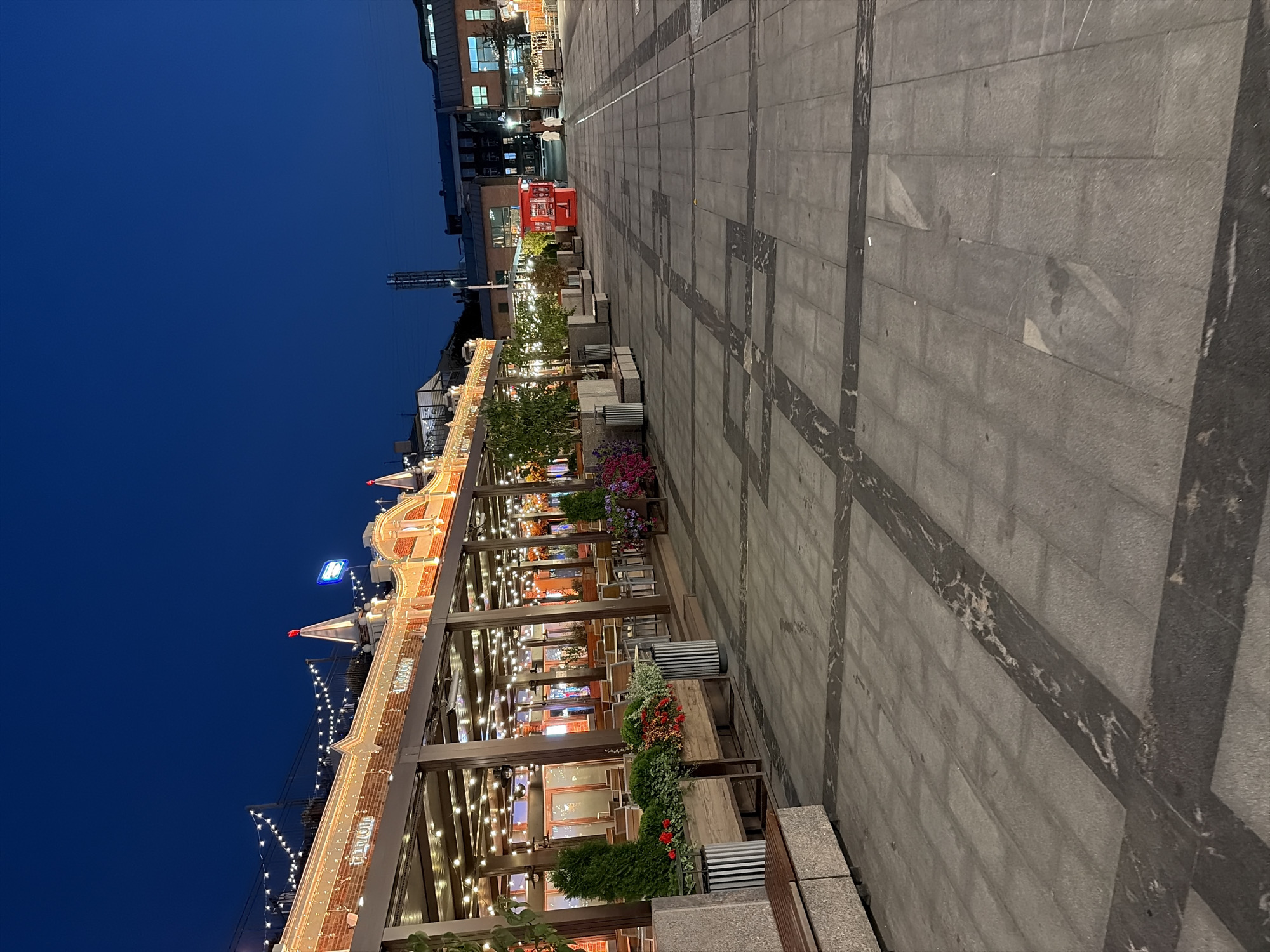The image captures an outdoor seating area of a restaurant or cafe at dusk in Moscow, Russia. The establishment features a covered patio with numerous tables and chairs, illuminated by strings of lights and interior lighting. Several planters with greenery and flowers are positioned along the edge of the patio. Adjacent to the seating area is a paved plaza with a geometric pattern formed by darker stone inlays. In the background, various buildings are visible, including a taller industrial-looking structure and a red kiosk. Two individuals, dressed in light-colored coats, are walking away from the red kiosk down the plaza. The sky is a deep blue, indicating it is nighttime.