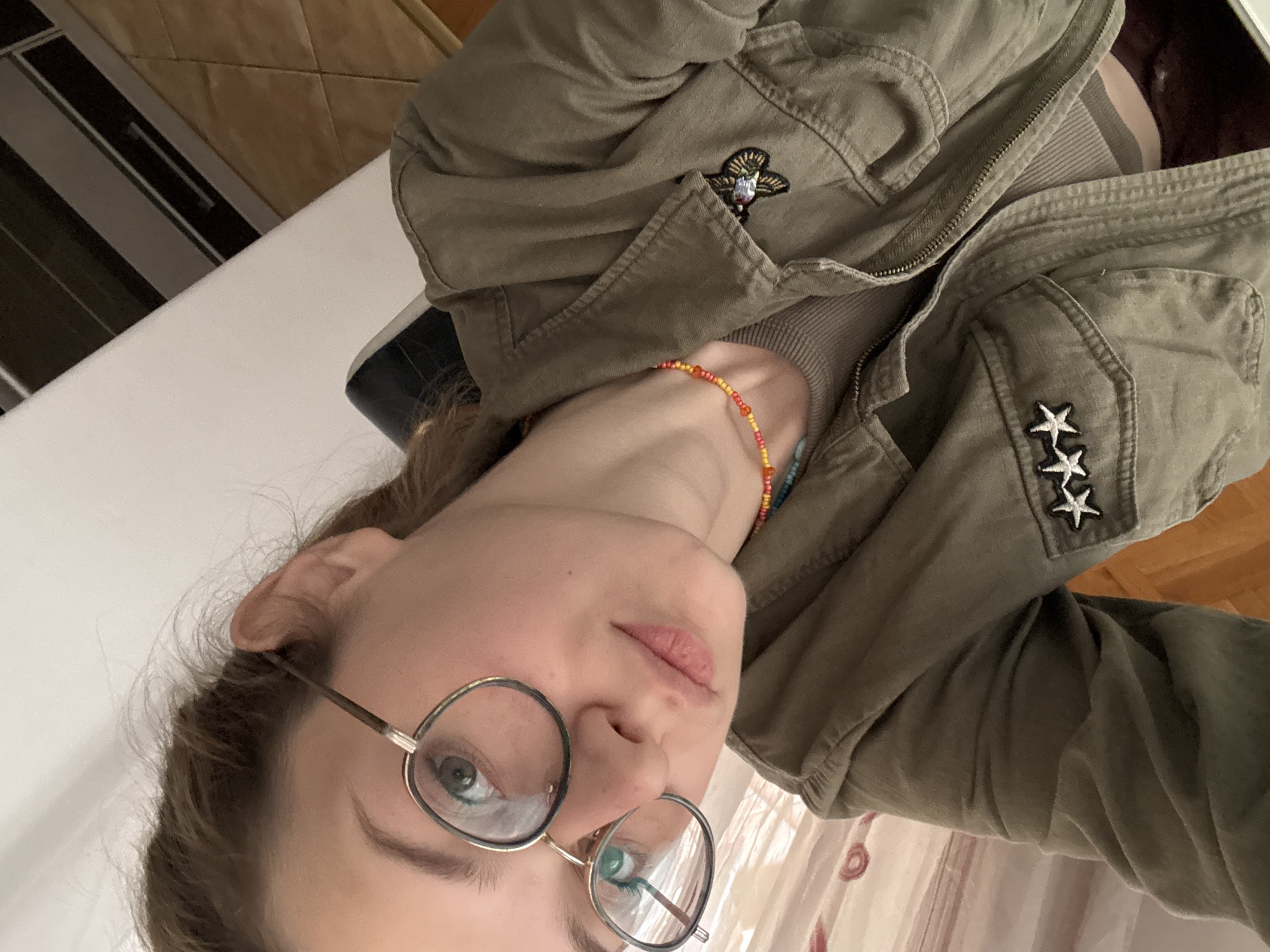 A woman is shown in a close-up, angled selfie. She wears round, wire-rimmed glasses and an olive-green jacket with decorative pins and patches, including stars and a stylized emblem. A colorful beaded necklace is visible. The background includes a glimpse of a white surface and a wood floor. The location is unknown and there is no evidence that this image was taken in Kragujevac, Serbia.