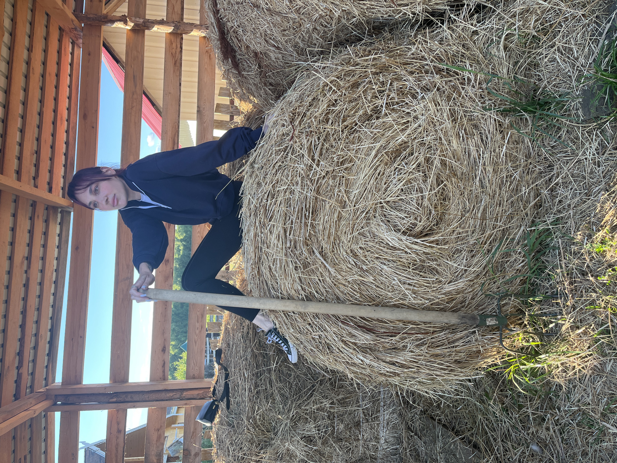 A young woman with dark hair is seated on a large bale of hay inside a rustic wooden structure. She is wearing a dark blue hooded sweatshirt and black leggings, with black Converse sneakers on her feet. She is holding a pitchfork upright in front of her. The structure appears to be an open-sided barn or shelter, with a wooden roof and slatted walls. In the background, there are glimpses of houses with red roofs and green trees, suggesting a rural or semi-rural setting. The ground around the hay bales is covered with straw and some green grass. The lighting suggests it is daytime, with sunlight casting shadows. The overall scene depicts a moment of rest or pause in an agricultural or outdoor environment. The image was taken in the village of Vybolovo, Russia.