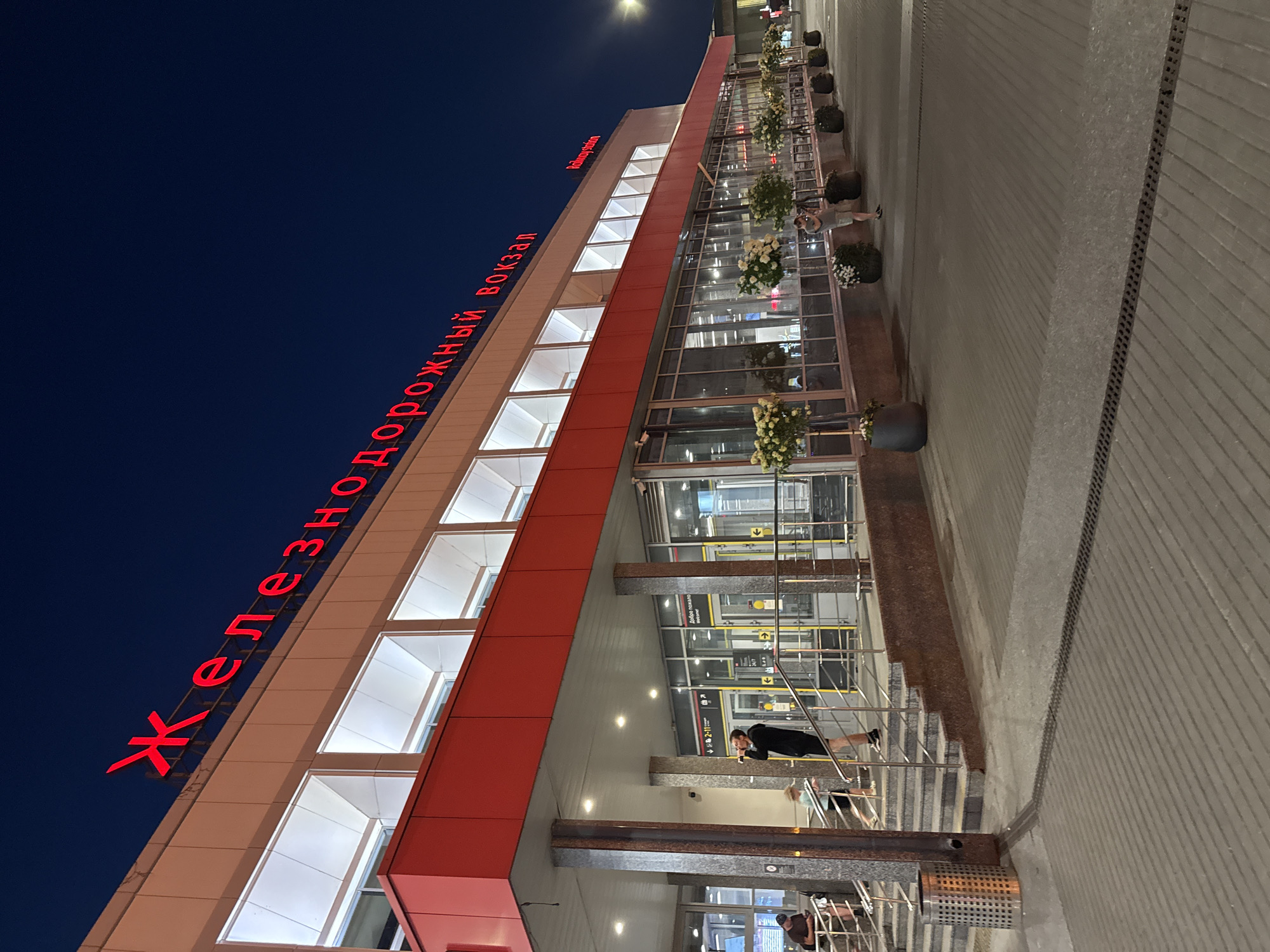 This is a low-angle, outdoor, eye-level shot of a modern railway station at night. The building's facade is predominantly white with a prominent red band running across the top and a larger red section above the entrance. Large, illuminated rectangular windows are visible on the upper level. The words "Железнодорожный вокзал" (Railway Station) are illuminated in red neon letters across the top of the building. To the right, smaller red neon letters spell out "Railway station".
The entrance area features a wide set of stairs leading up to automatic doors. Several people are present. On the stairs, a man in black shorts and a black hooded jacket is standing and holding a phone to his ear, appearing to be speaking. Further up the stairs, a child in a light-colored t-shirt and shorts is running. On the left side of the stairs, near a metal trash receptacle, two individuals are seated, one on the steps and another on a lower level.
The ground in front of the station is paved with dark gray interlocking bricks laid in a diagonal pattern. A drainage grate runs across the paving stones. Along the front of the building, a row of large, dark gray planters contains trees with white floral clusters. A man in a light-colored t-shirt and shorts is standing near one of these planters, holding a camera and appearing to take a photograph. The sky is a deep, dark blue. The overall lighting suggests artificial illumination from the building and surrounding streetlights, creating a twilight or nighttime ambiance.