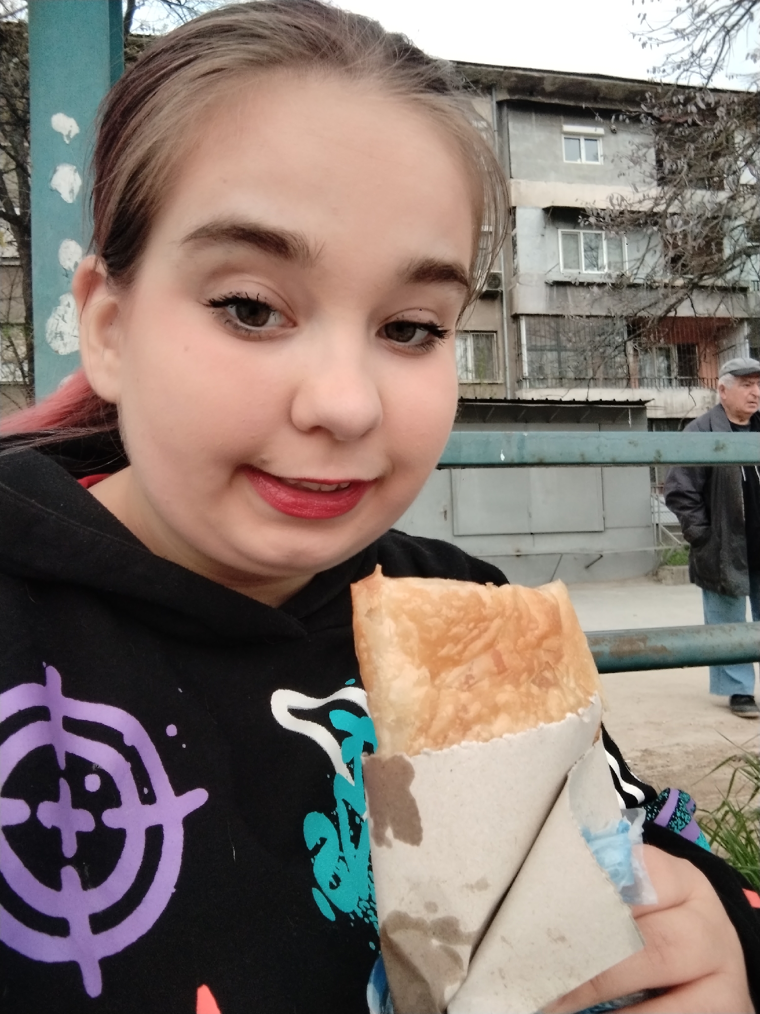 A young woman with fair skin, dark eyebrows, and red lipstick is holding a pastry in a paper wrapper. She is wearing a black hoodie with colorful designs. The pastry is golden brown and appears to be freshly baked. In the background, an older man wearing a grey cap and dark coat is walking on a sidewalk with a low metal fence. Behind him is a multi-story building with windows and balconies. The scene is an outdoor setting, likely a street in a city. Given the buildings and style, the location is most likely in Plovdiv, Bulgaria.